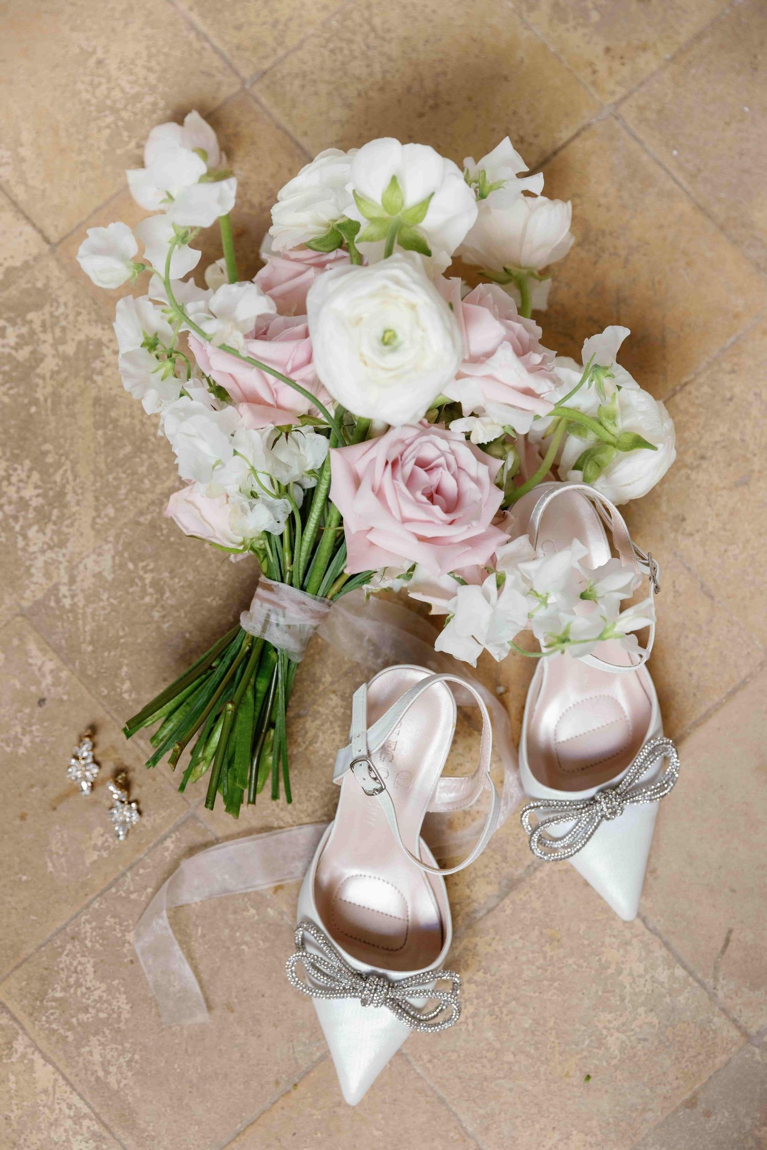 Bridal flat lay with blush pink and white flower bouquet, crystal bow heels, and delicate earrings on stone floor.
