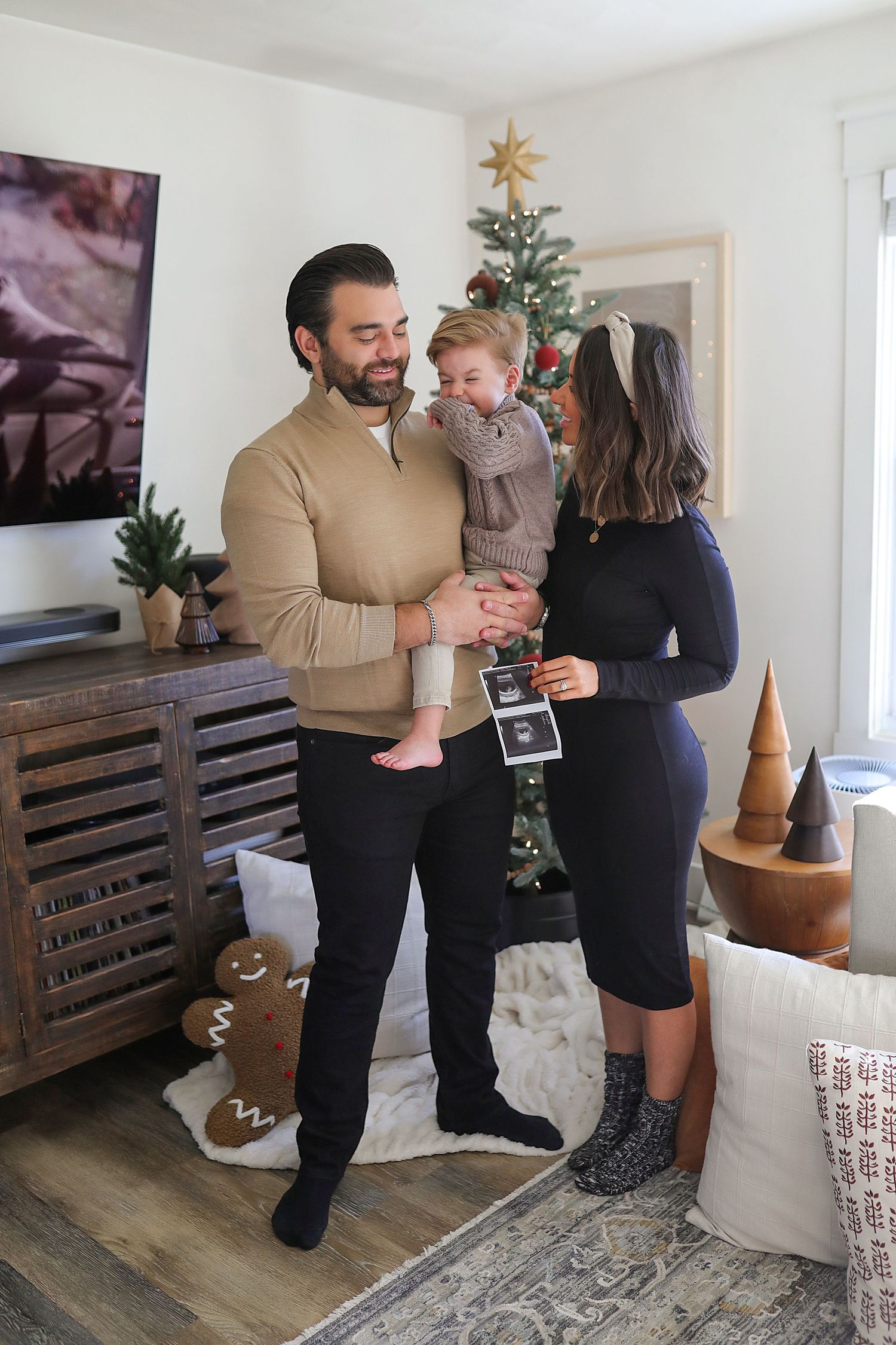 Family stands together by wooden pallet decorations and a Christmas tree in a bright modern living room interior.