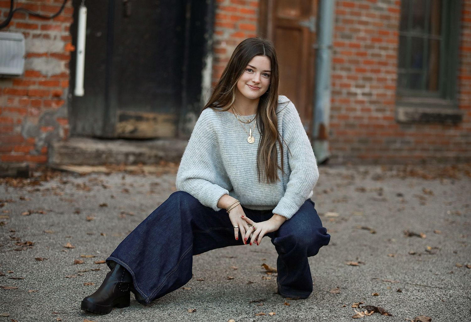Senior Photography in Chagrin Falls, OH. Person in a white sweater and wide-leg jeans sits casually on the ground against a brick building.