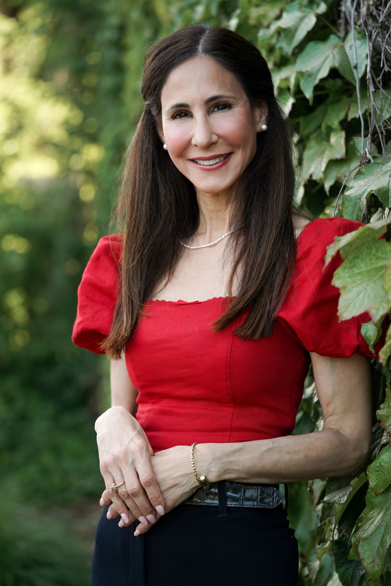 A person in a red crop top and dark pants poses outdoors against a backdrop of green ivy leaves in natural lighting.