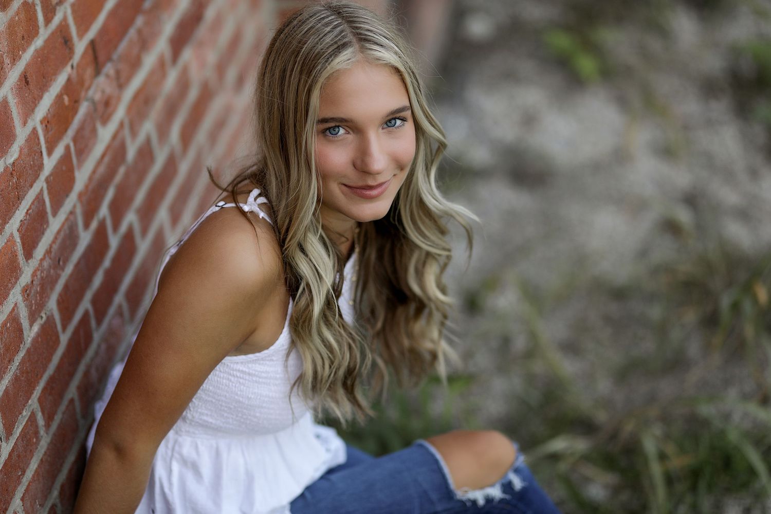 Woman in a white strapless dress stands against a rustic brick wall with vines in an outdoor setting.