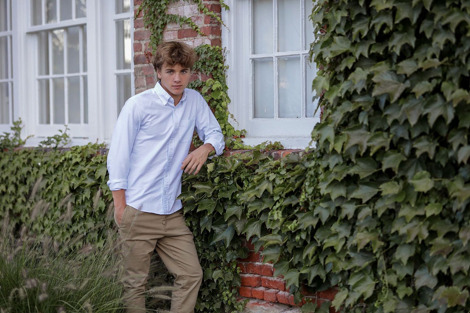 A person in a white button-down shirt and khaki pants leans against an ivy-covered brick wall near white windows.