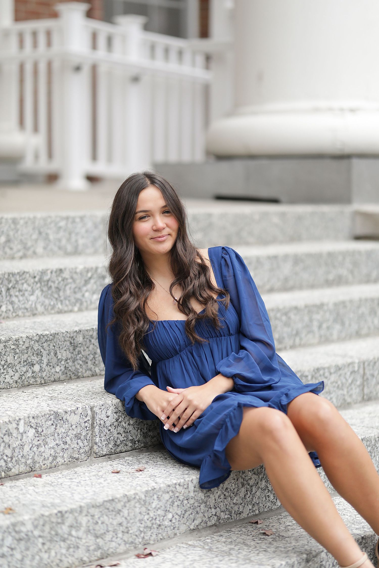 Young woman in flowing navy blue dress sitting on concrete steps on a sunny day.