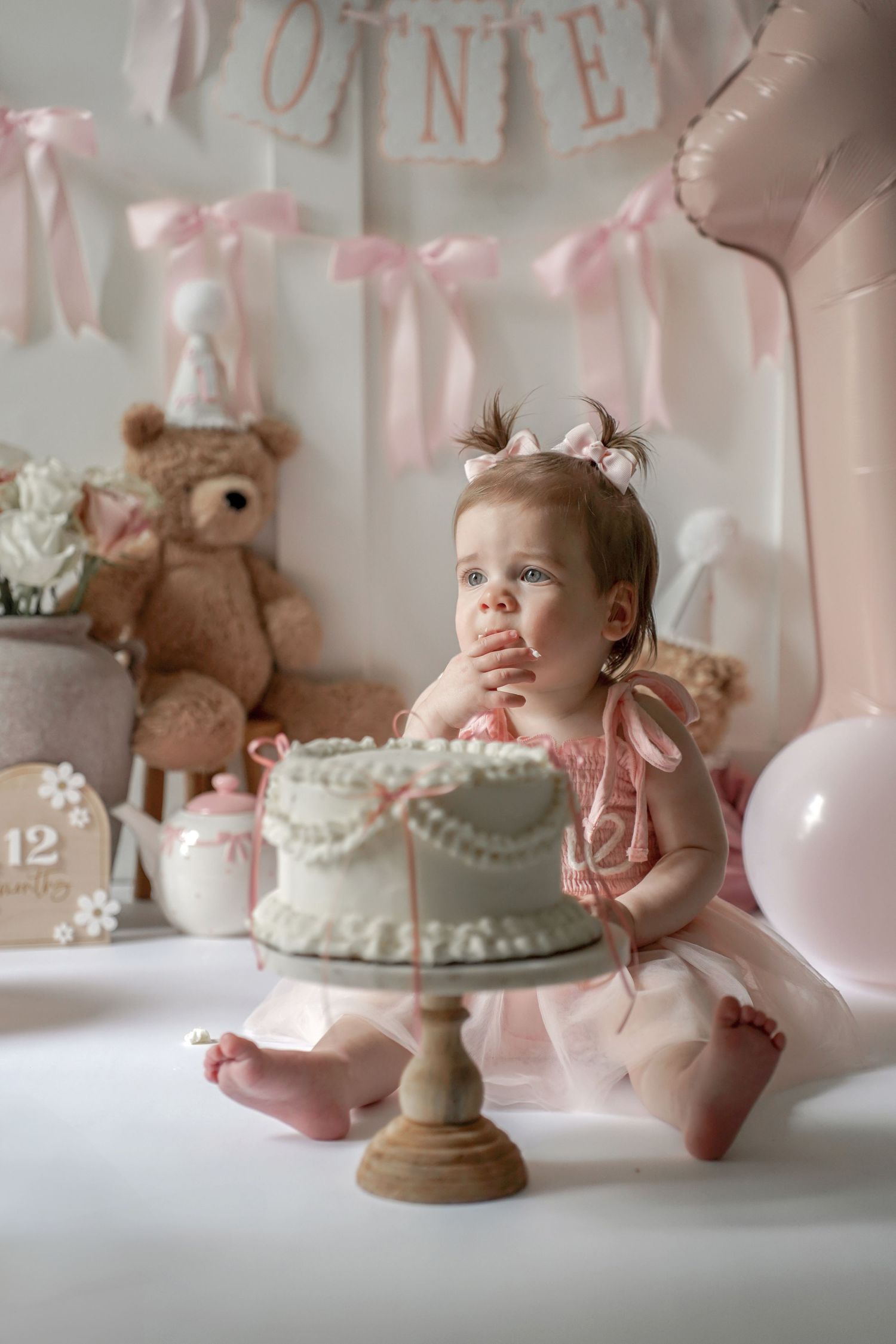 First birthday celebration setup with mint green cake on wooden pedestal stand, pink decorations and teddy bear in background.