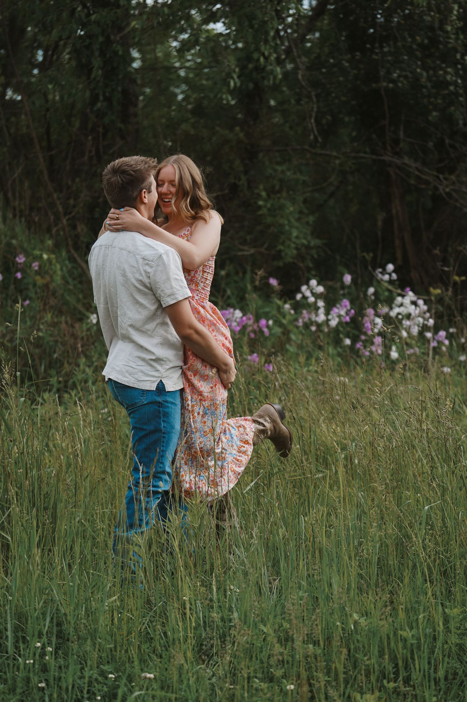 A romantic couple embraces in a meadow of tall grass and wildflowers on a summer evening.