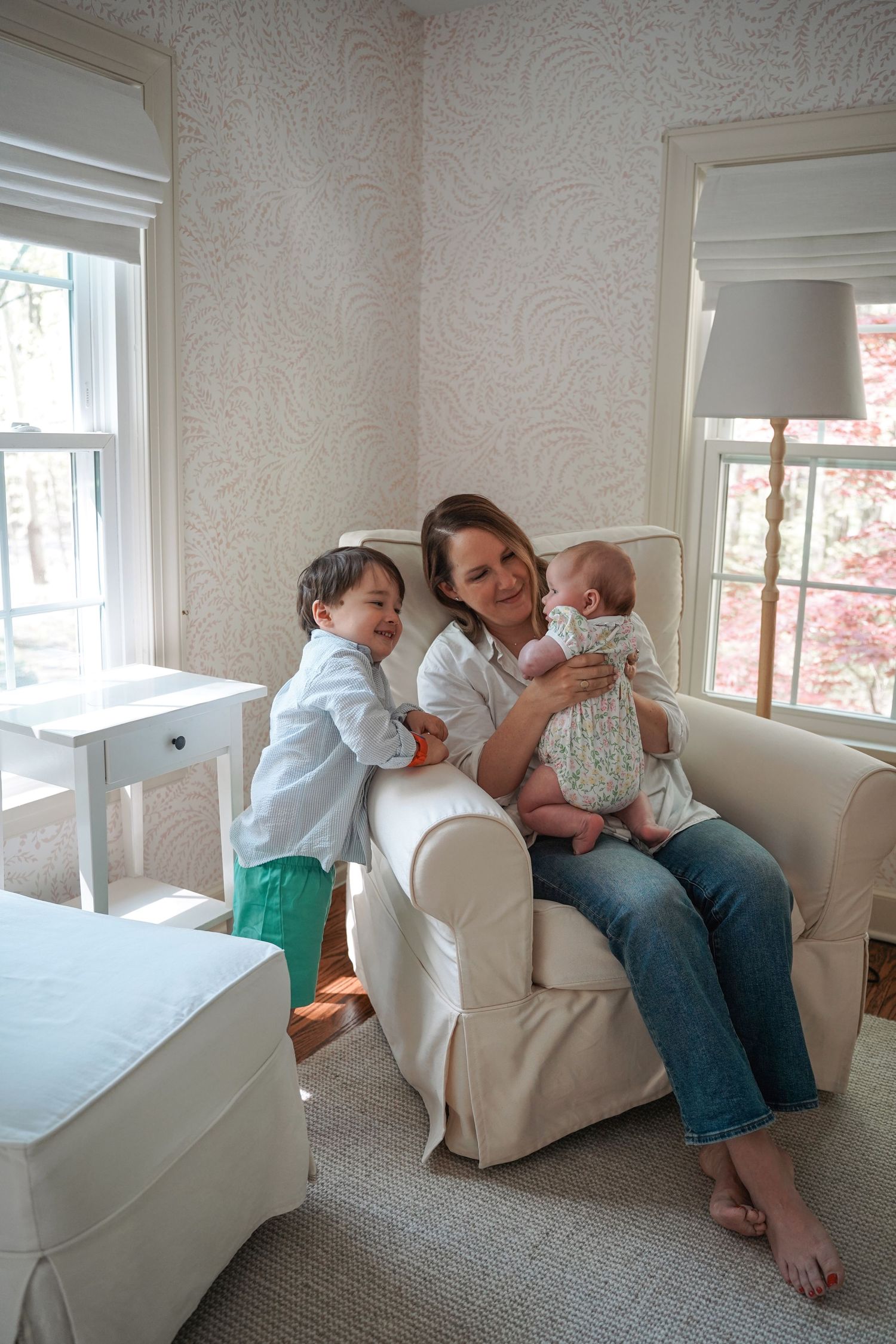 A cozy nursery scene with family members sharing a tender moment on a white armchair near sunlit windows.