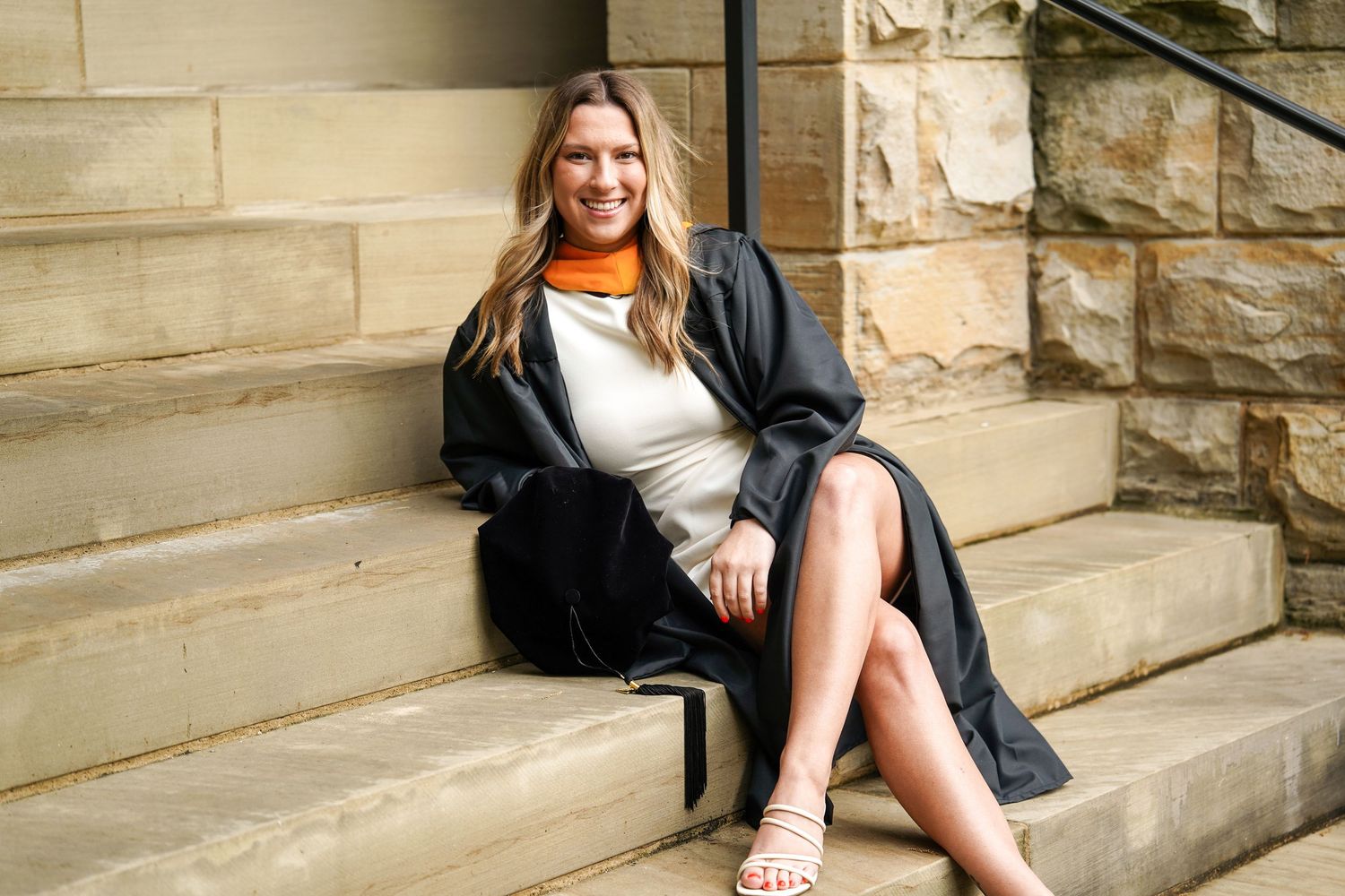 Someone wearing an orange turtleneck and grey jacket sits elegantly on stone steps against a textured wall.