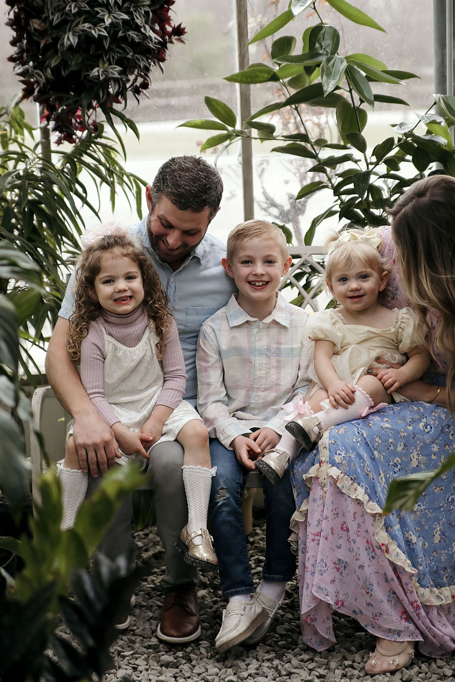 A family sits together in a lush greenhouse surrounded by green plants and foliage for an intimate portrait session.