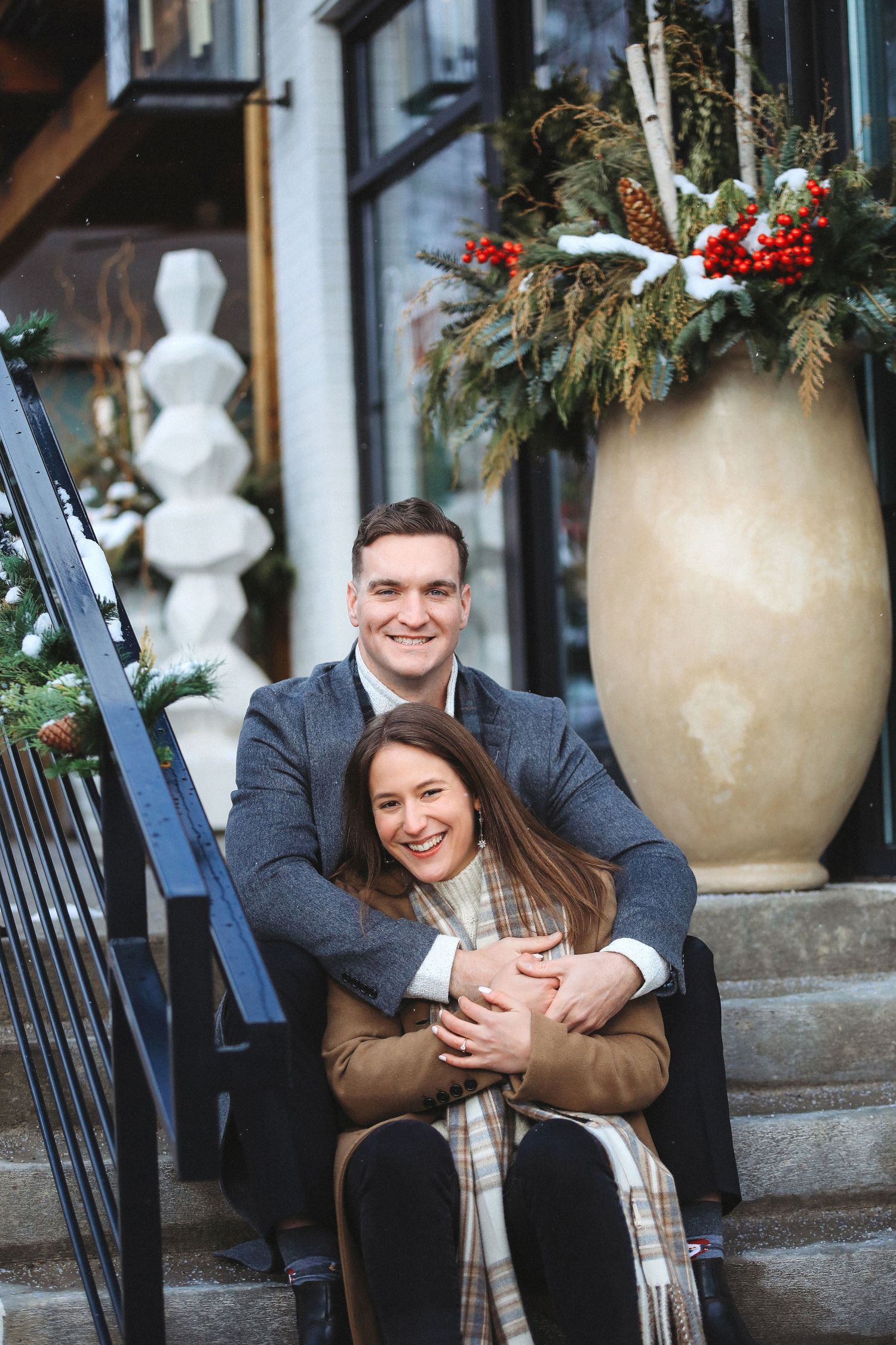 A couple shares a joyful embrace while sitting on outdoor steps decorated with holiday greenery and large urns.