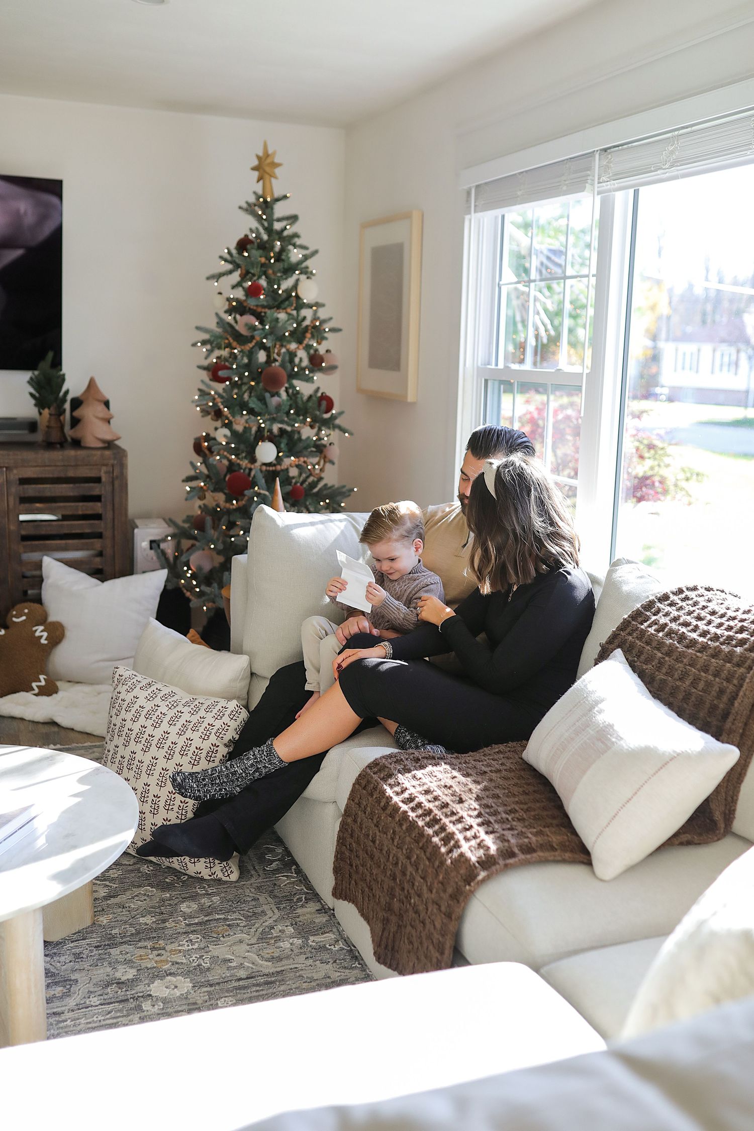 A cozy living room with a decorated Christmas tree, city view window, and figures relaxing on a cream colored couch.