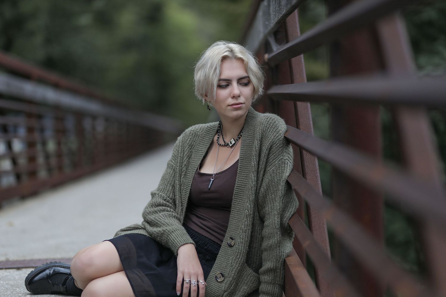 Person in olive cardigan and black dress sitting on rustic bridge railing during overcast day.