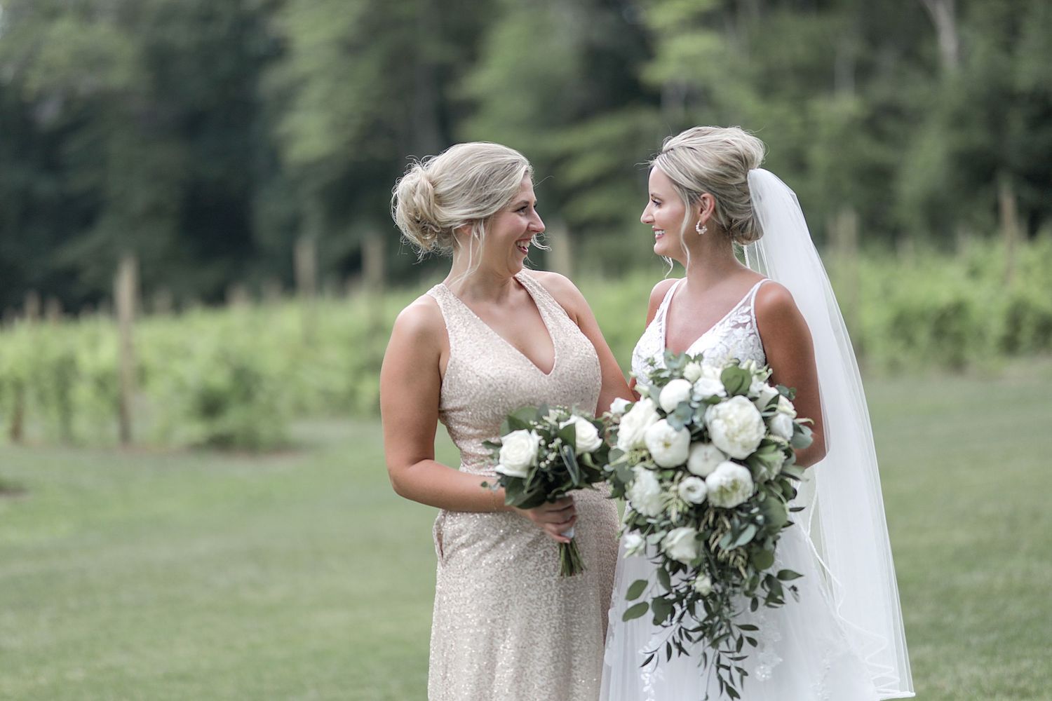 Two wedding attendants in light-colored dresses hold white bouquets on grassy lawn.