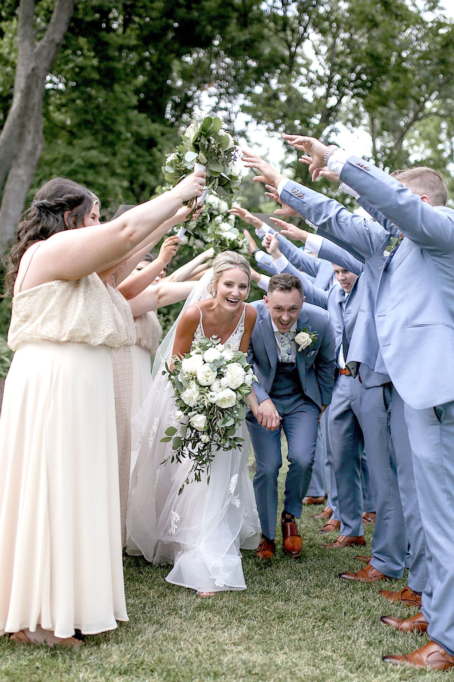 Wedding party in gray suits and blush dresses posing playfully on a grassy lawn.