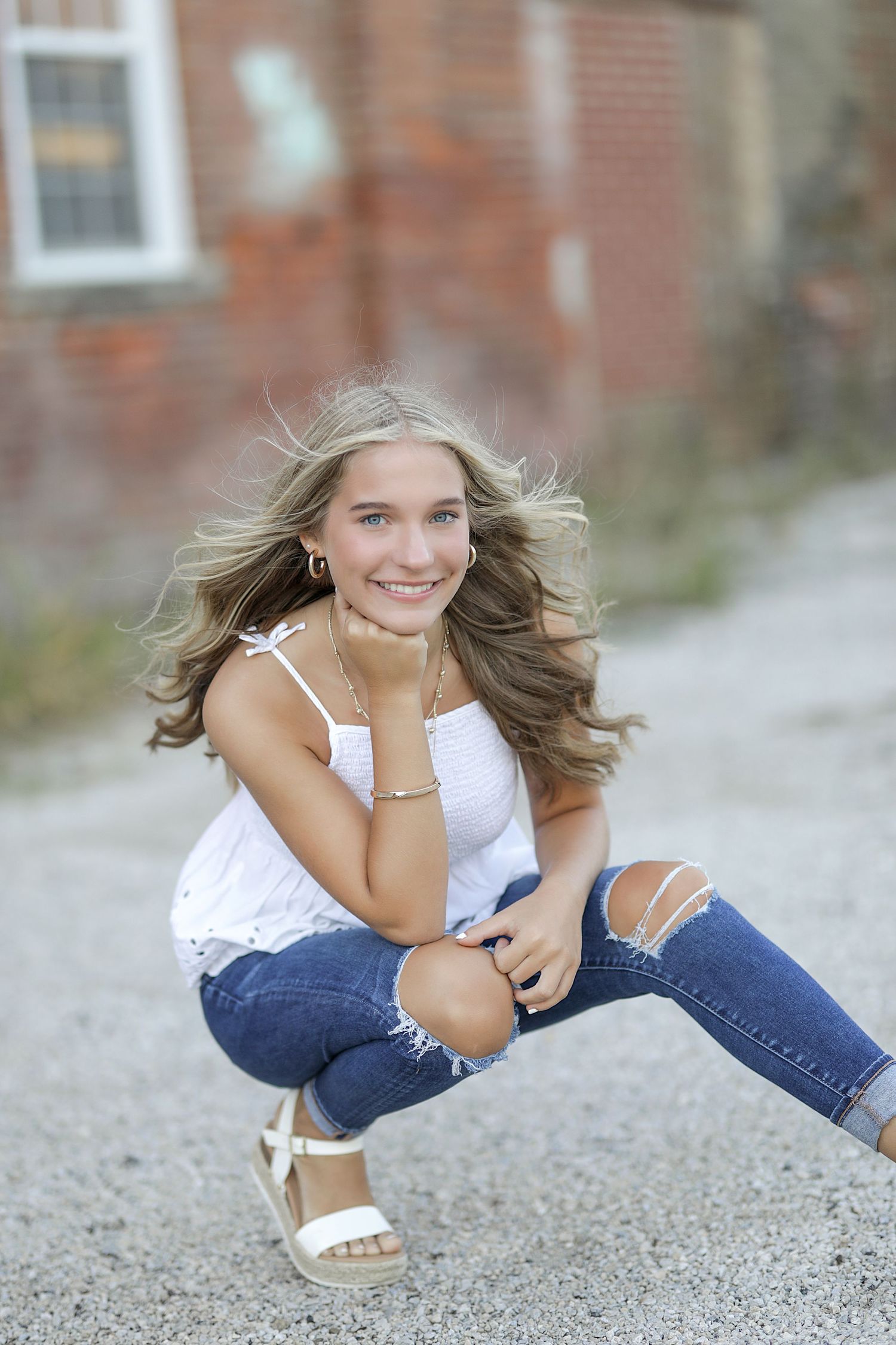 A person in ripped jeans and white top crouches outdoors with wind-blown hair and a bright smile.