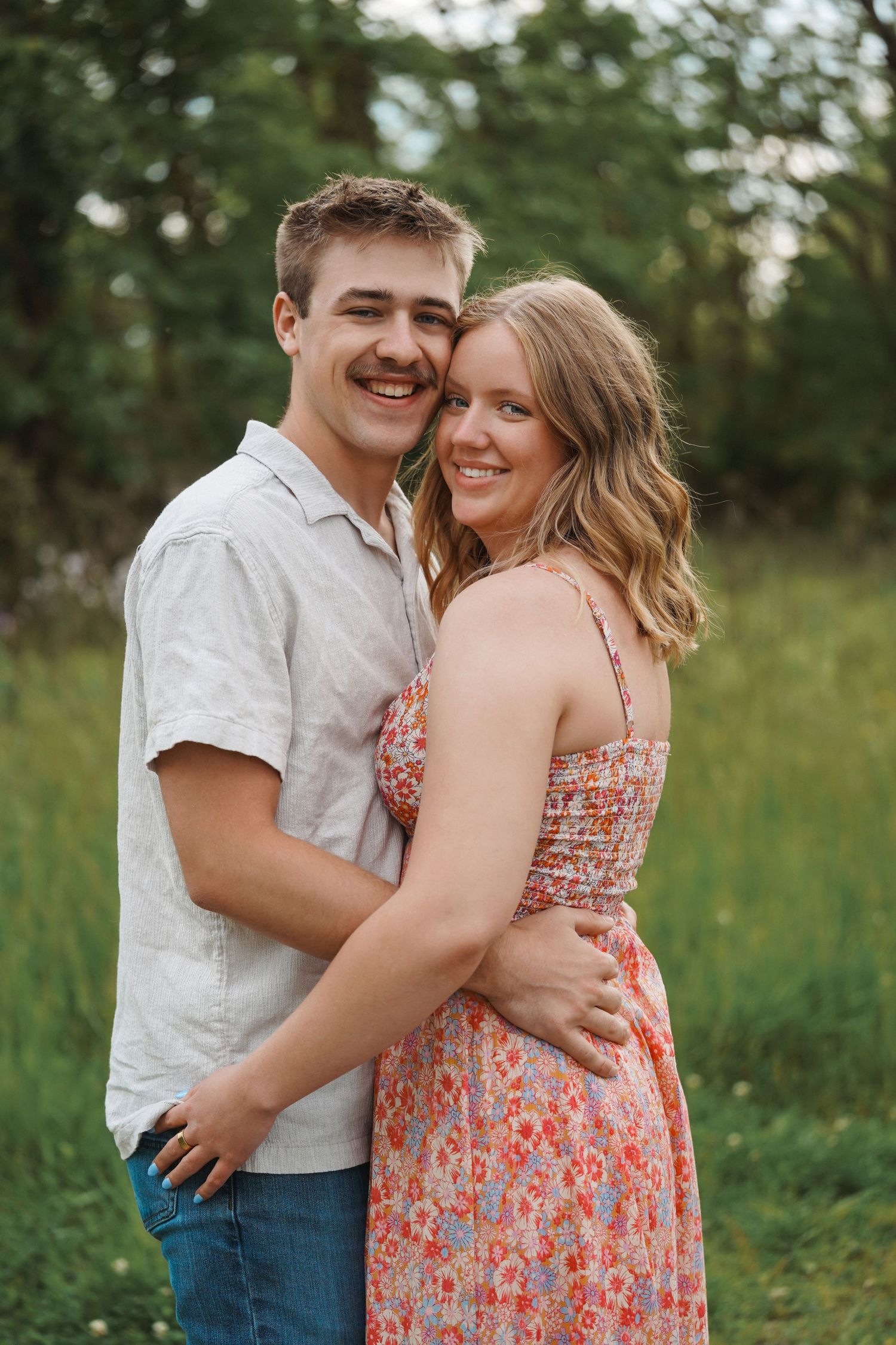 A couple embraces and laughs together during a romantic photoshoot in a sunny meadow with green foliage.