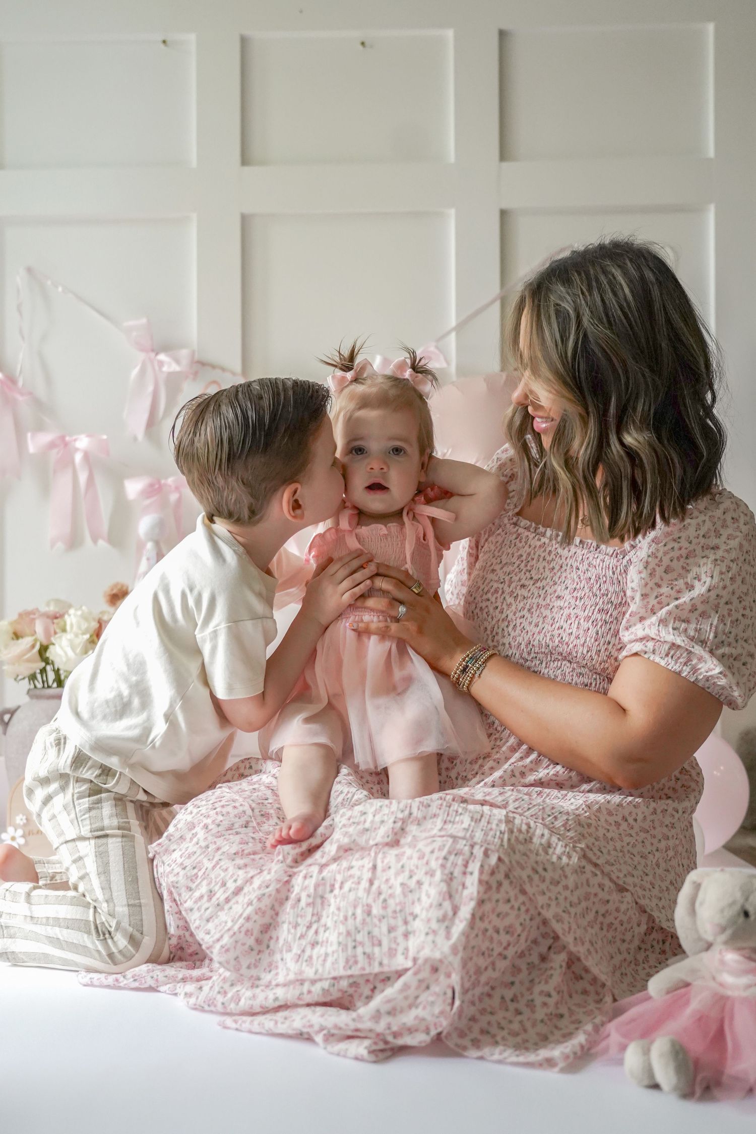 Sweet family moment captured on white bed with pink decor and ribbons in a light filled room.