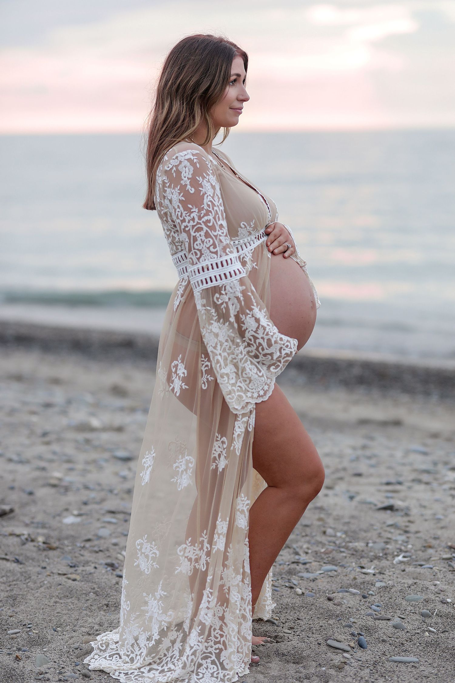 A pregnant woman in a sheer lace robe poses on the beach at sunset.