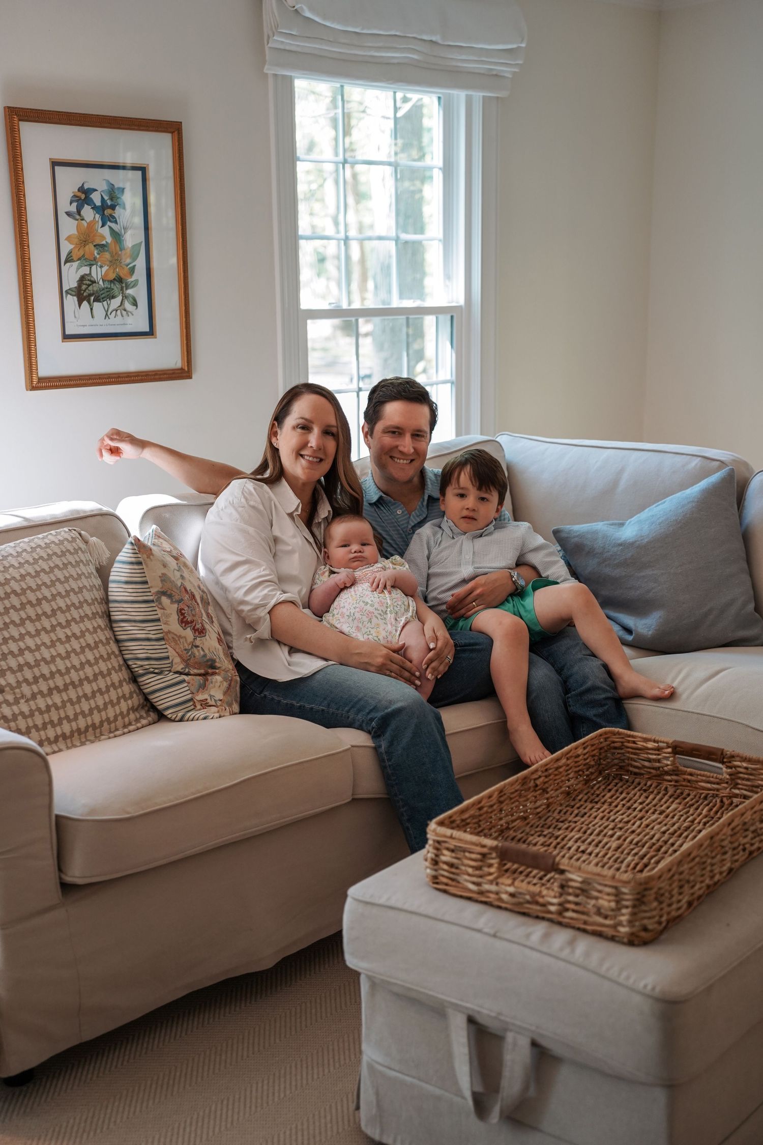 A family relaxes together on a light gray couch in a cozy living room with natural light coming through a window.