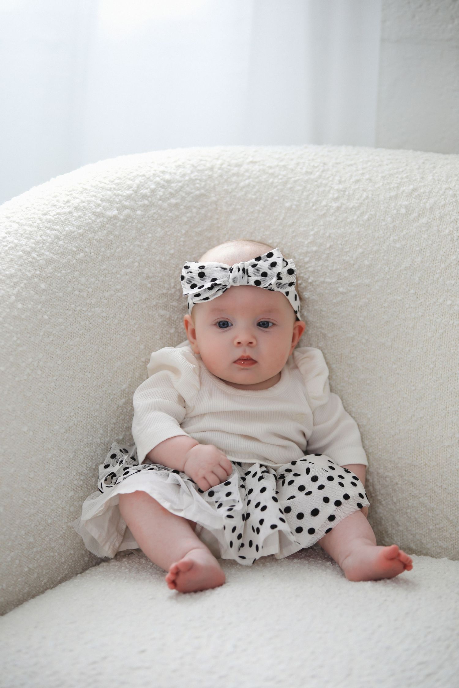 Baby wearing white onesie and polka dot bow lies on plush white couch in series of matching photos.