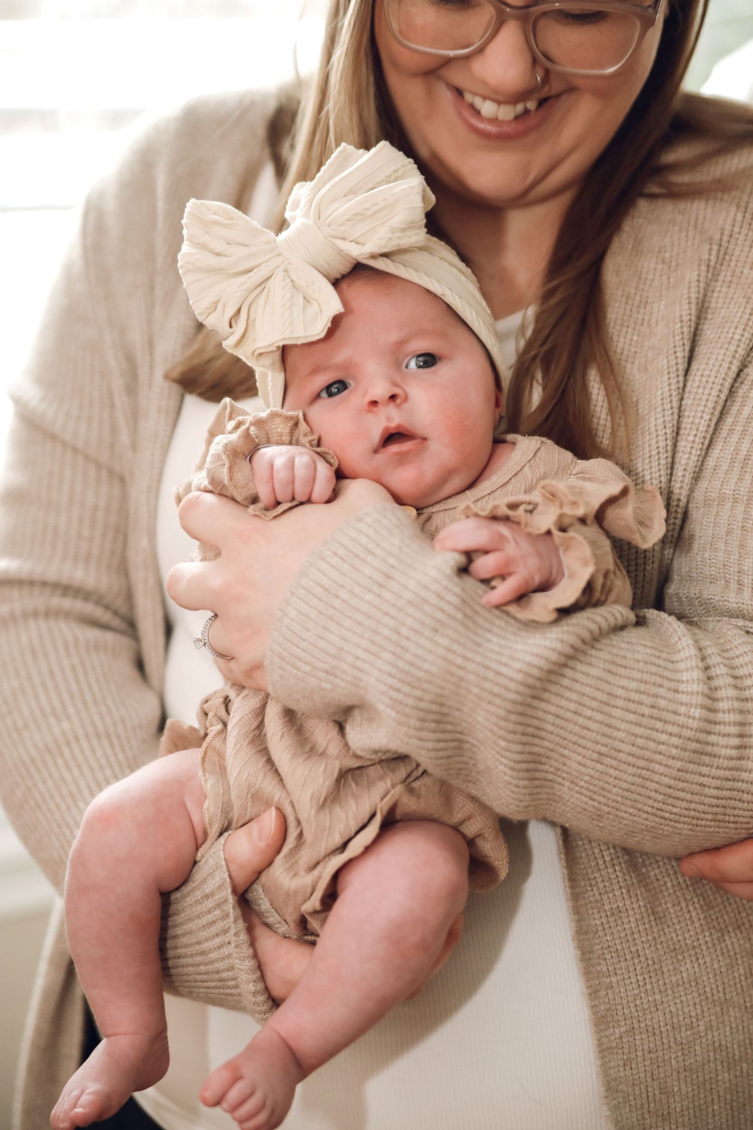 A baby wearing a beige ruffled outfit and bow headband is held in someone's arms in a warm-toned portrait series.
