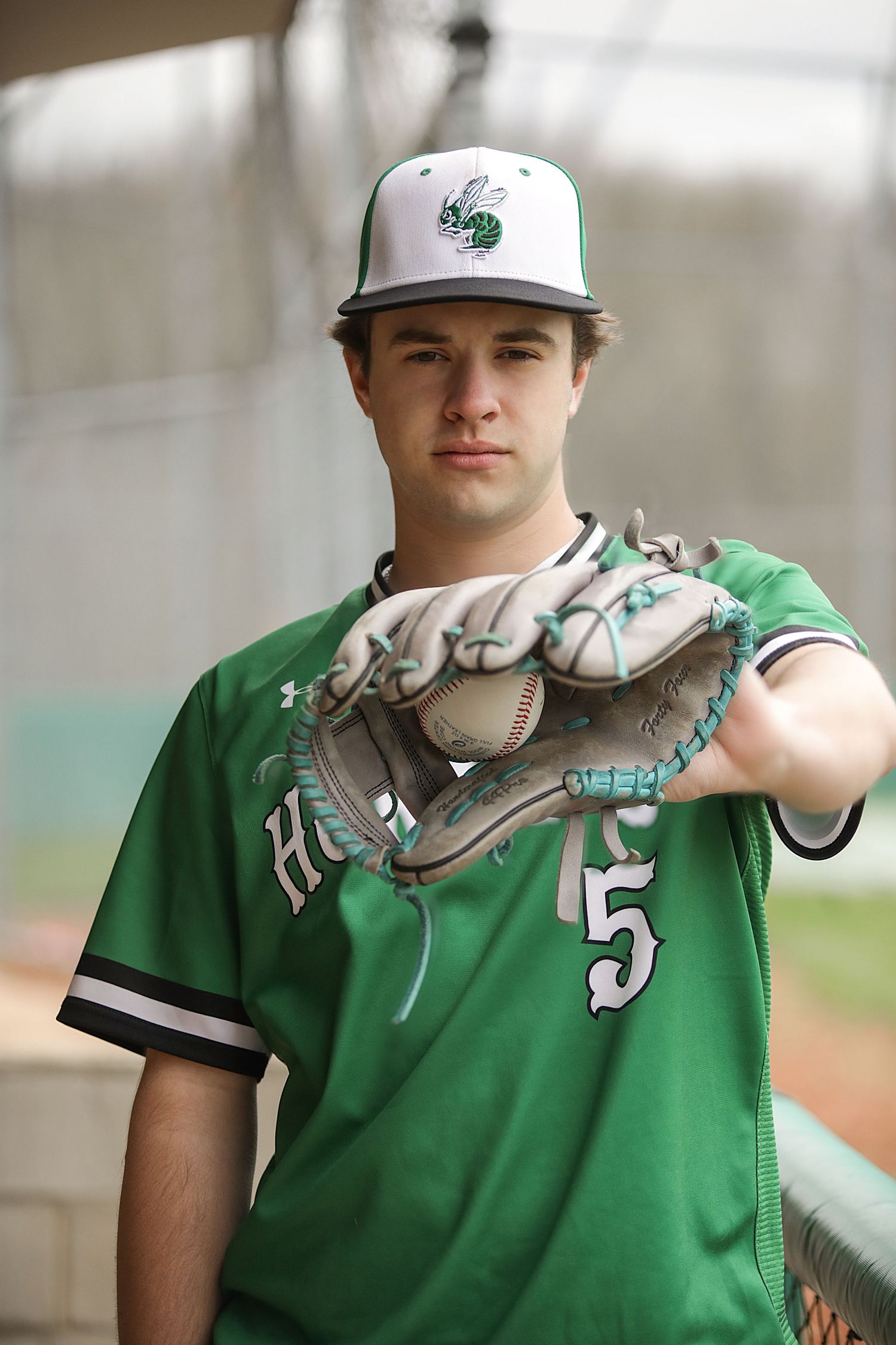 Baseball player in green jersey and white cap holds a baseball glove while standing on field.