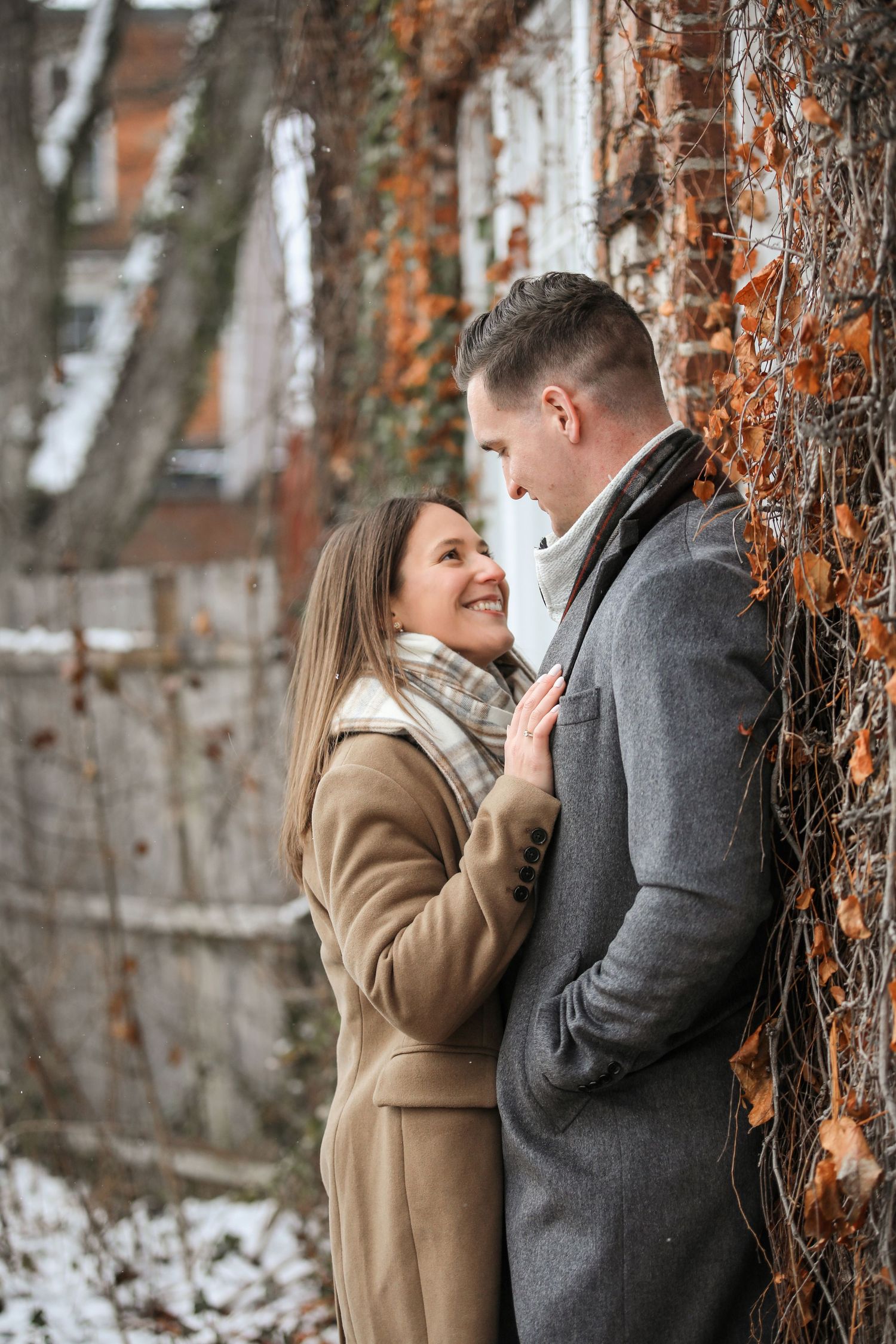 A romantic winter couple in wool coats share an intimate moment against a brick wall with snow and autumn leaves.