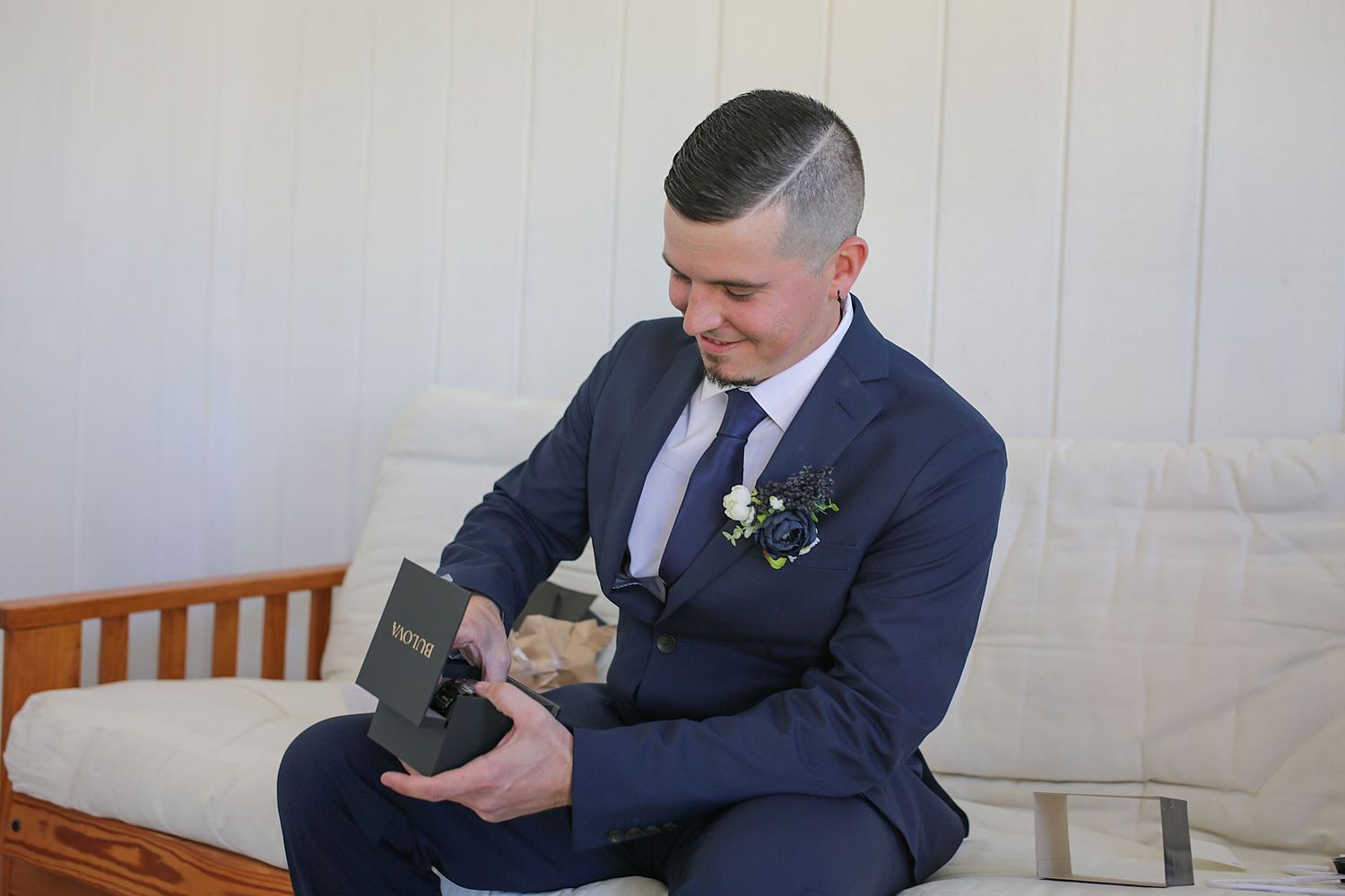 Someone in a navy blue suit adjusts their tie while sitting on a wooden bench before a formal event.