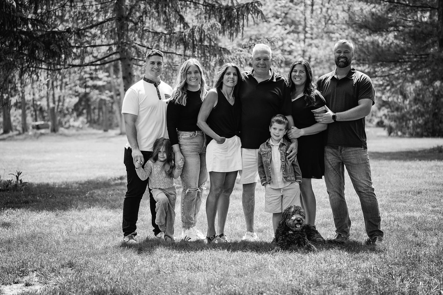 A black and white outdoor portrait of a multi-generational family group standing together with evergreen trees in background.