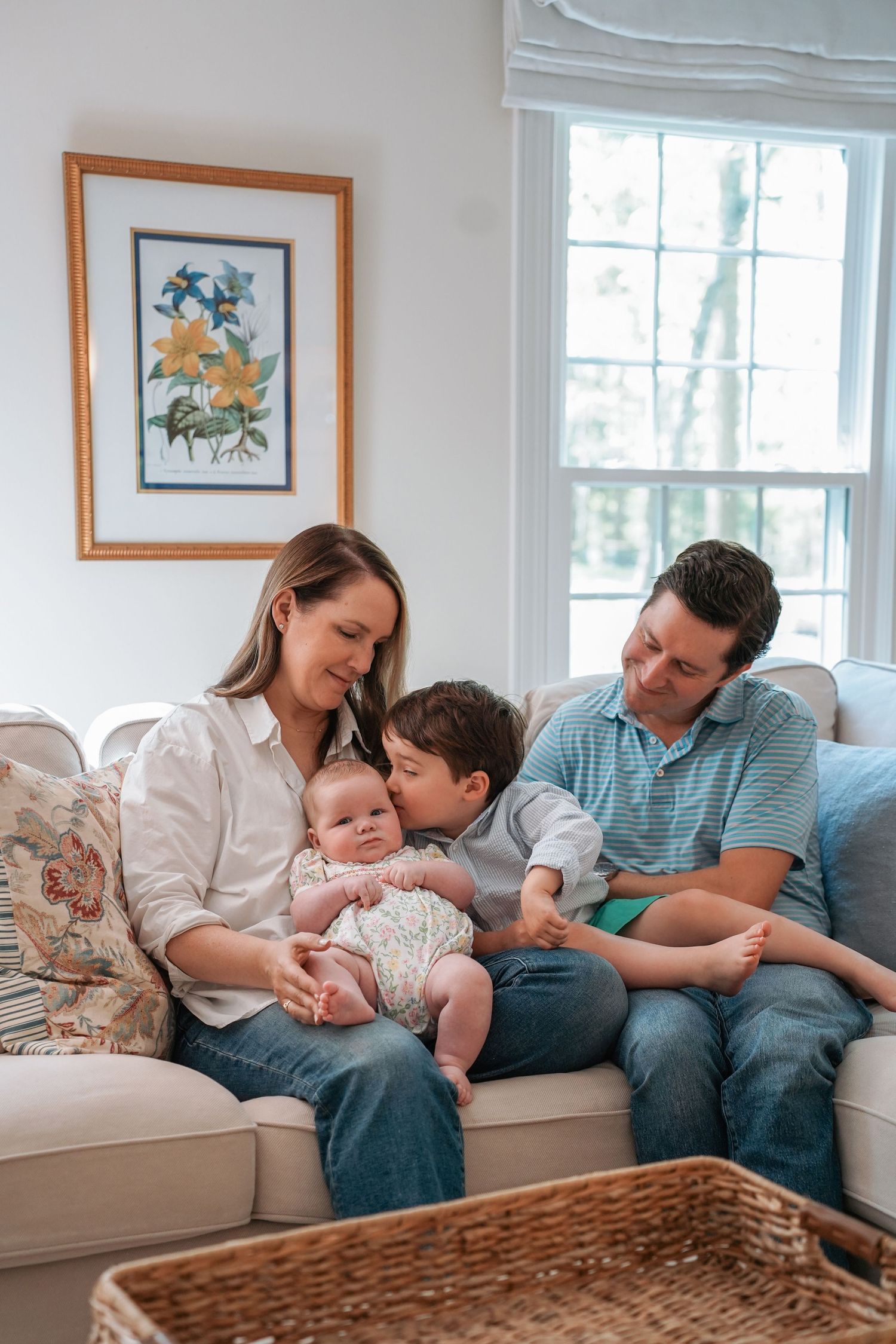 A family relaxes together on a beige couch in a bright living room with a floral print on the wall.