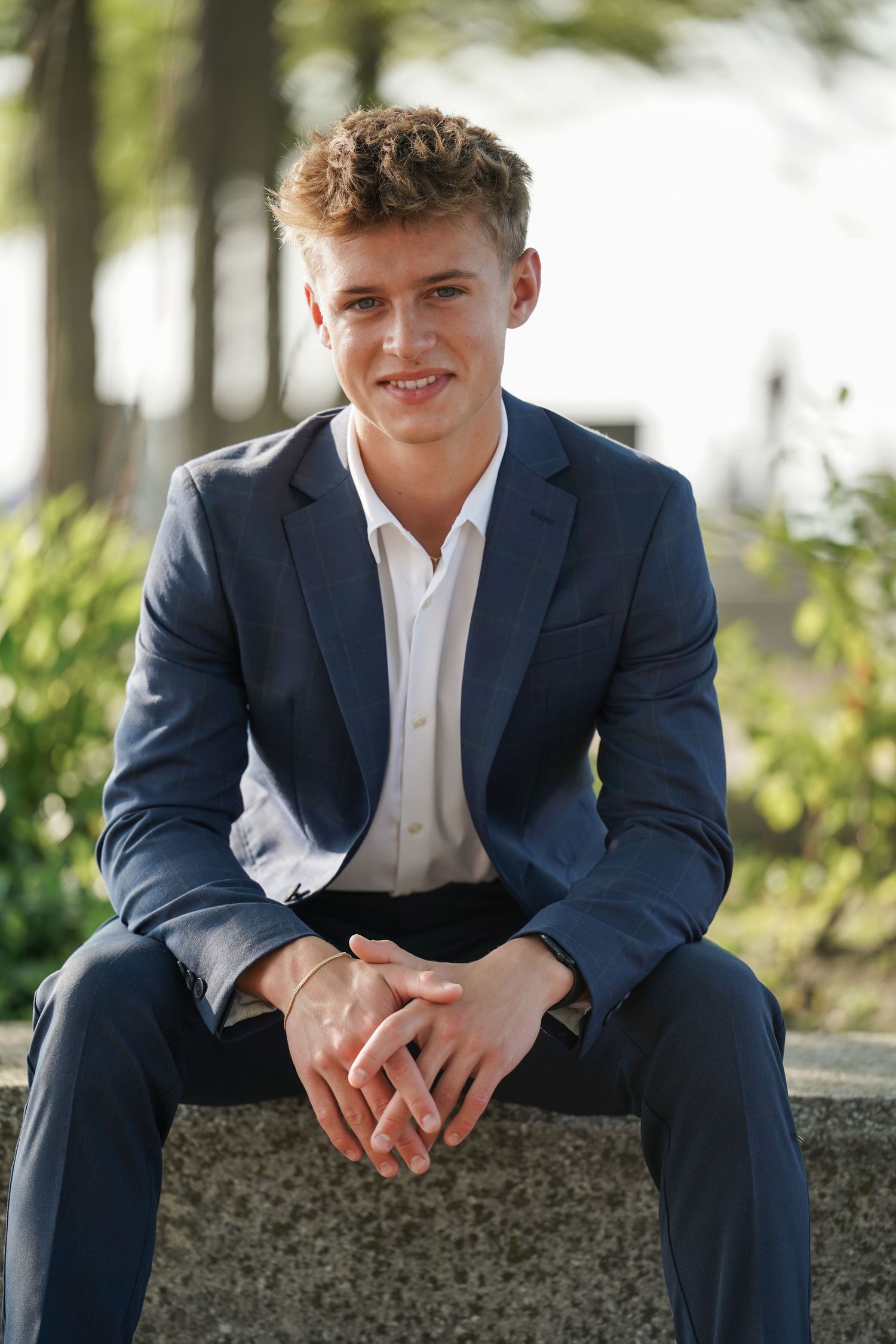A person wearing a navy suit and white dress shirt sits casually on a stone wall in an outdoor setting.