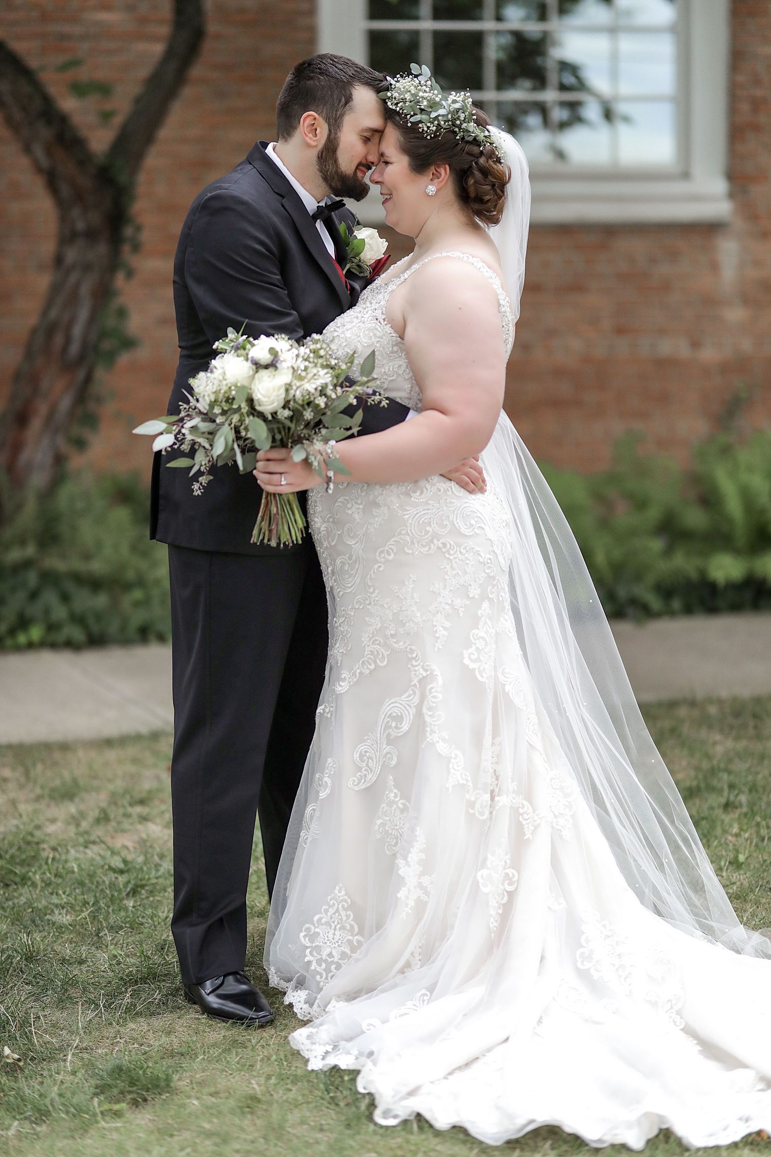 Newlyweds share intimate moment in garden setting with bride in lace gown holding white bouquet and groom in black tuxedo.