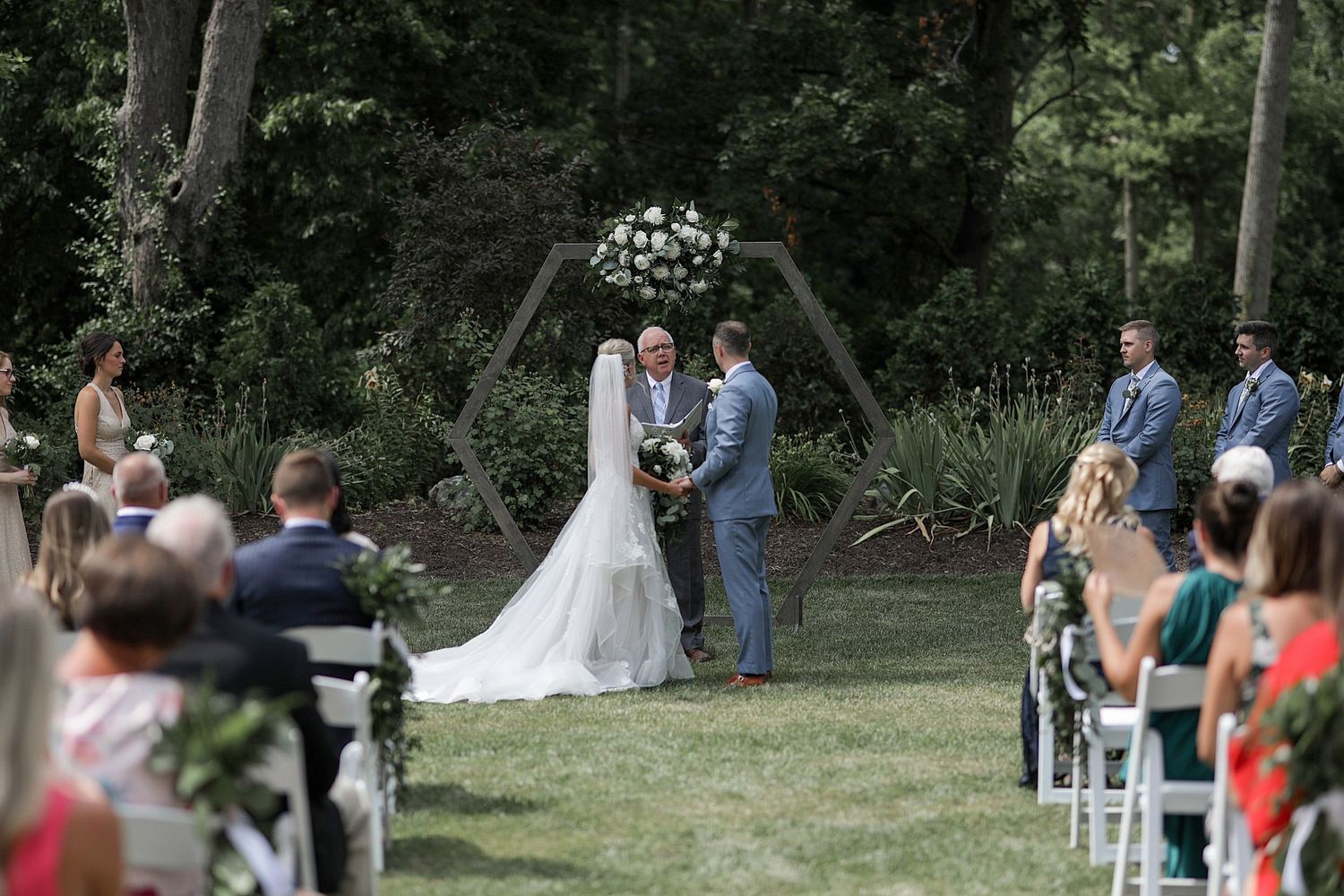 An outdoor wedding ceremony under a floral arch with guests seated on white chairs in a garden setting.