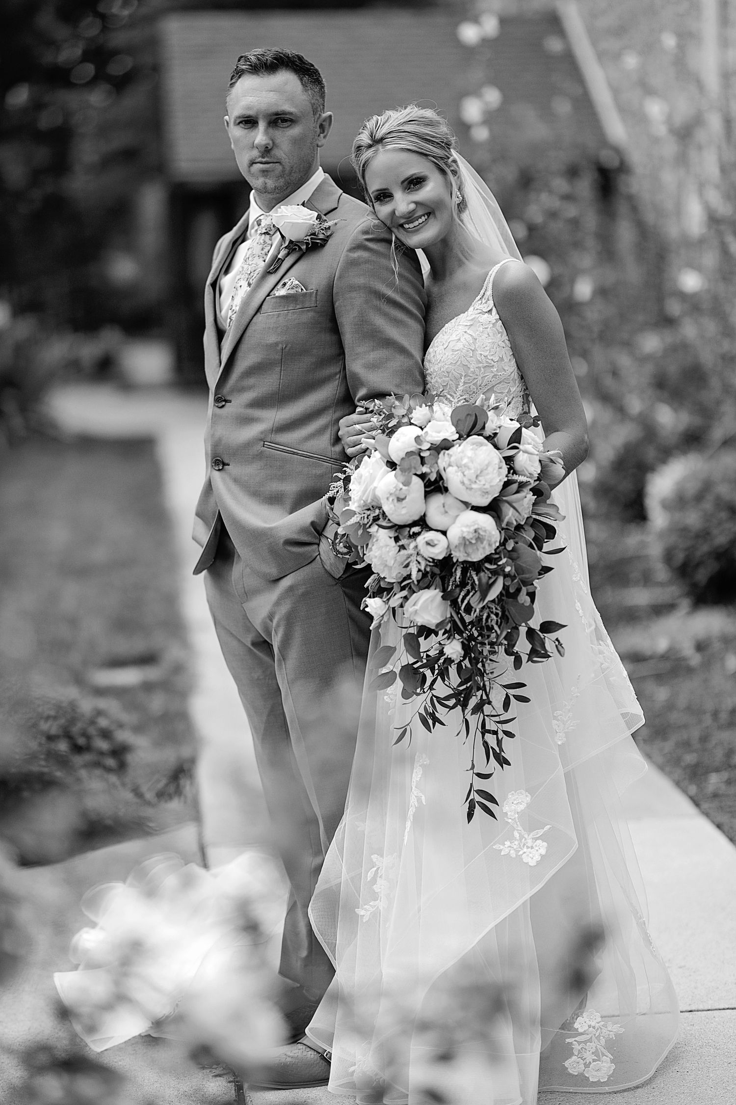 A black and white portrait of newlyweds with a lush floral bouquet in an outdoor setting.