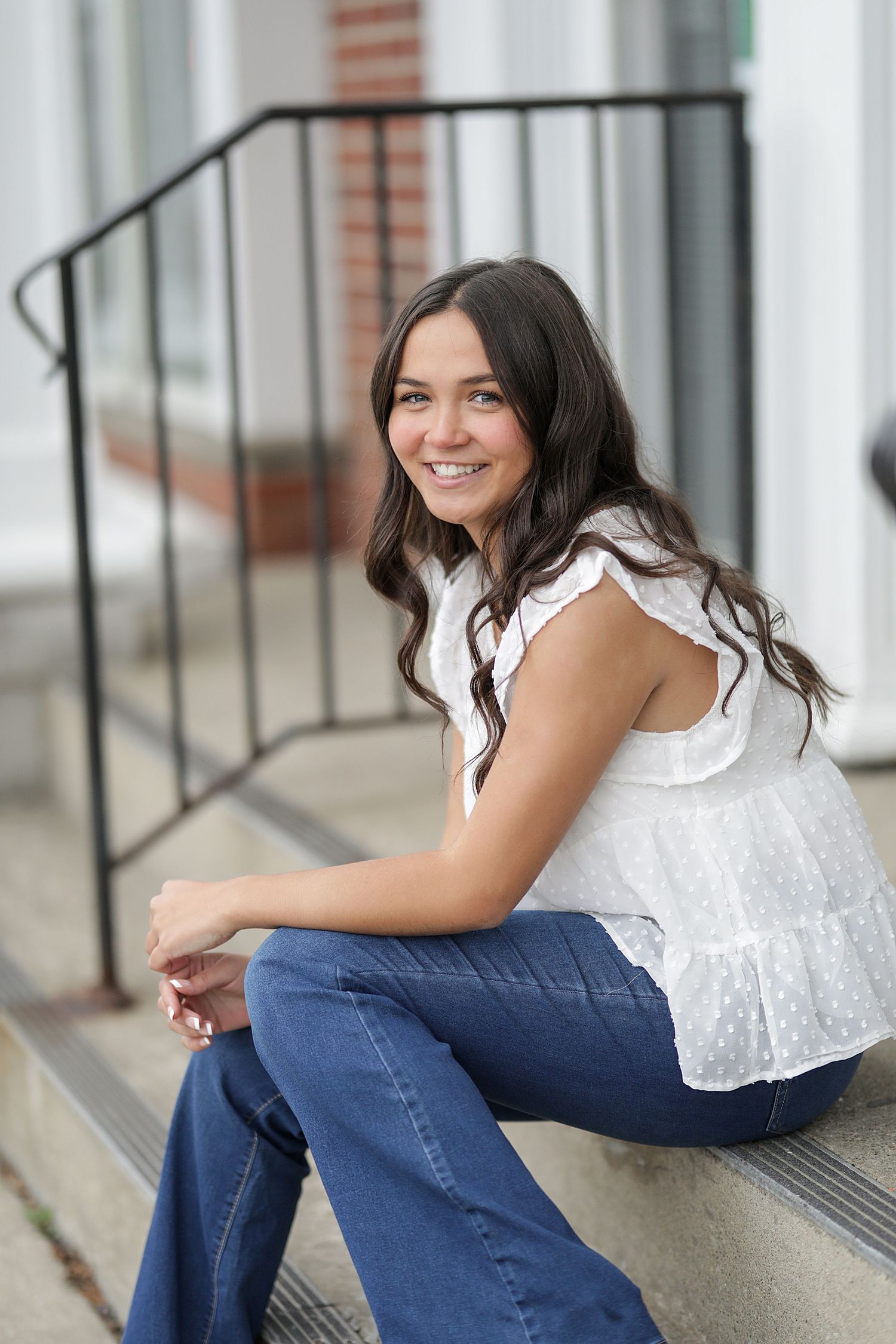 A woman in a white lace top and blue jeans sits casually on outdoor concrete steps with long dark hair and a bright smile.
