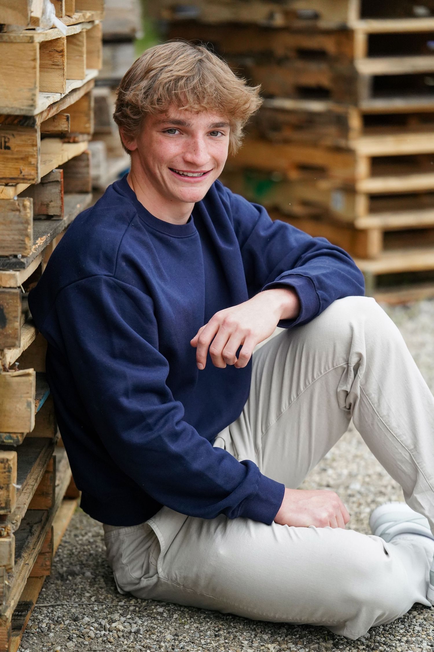 A person in a navy blue sweater and light gray pants sits casually on gravel near wooden pallets.