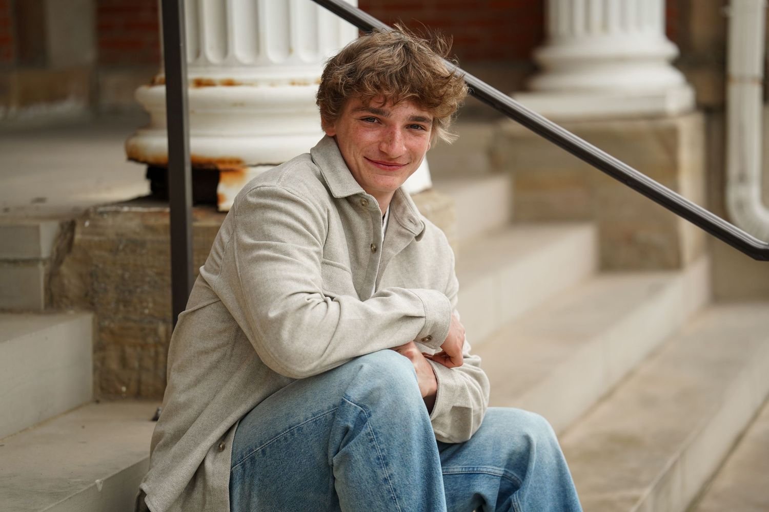 A person in a tan jacket sits casually on outdoor stone steps, wearing blue jeans and smiling at the camera.