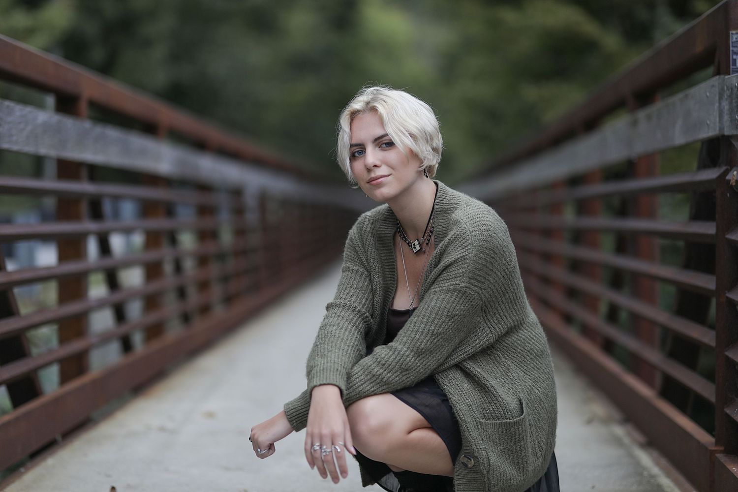 Person in olive green cardigan sitting on a metal bridge with blurred forest background.