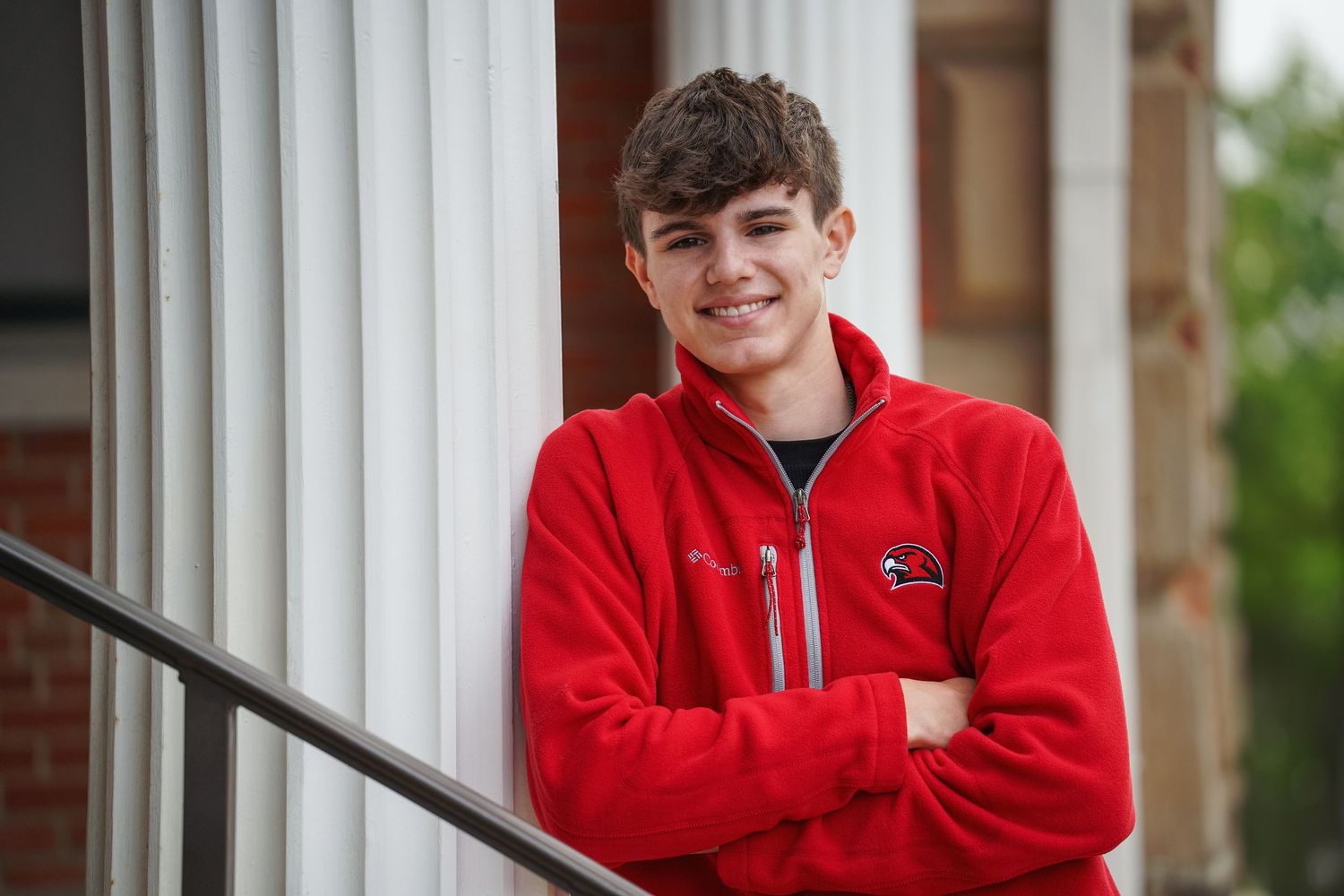 Person wearing a red hoodie smiles while sitting near white pillars at a brick building entrance.