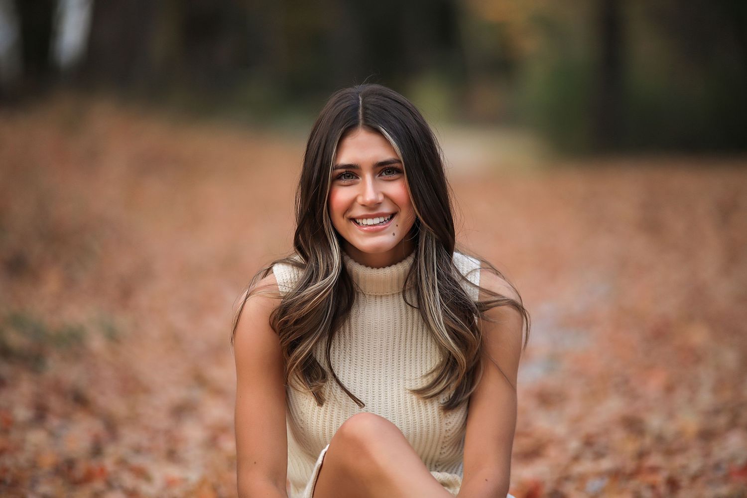 A portrait of someone sitting outdoors among fallen autumn leaves wearing a beige ribbed sweater.