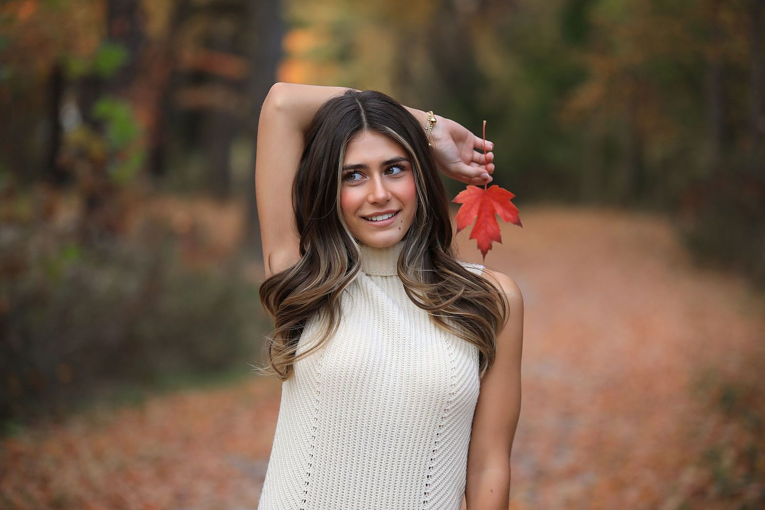 Woman in cream sweater walking through autumn forest path with fallen leaves in warm natural lighting.