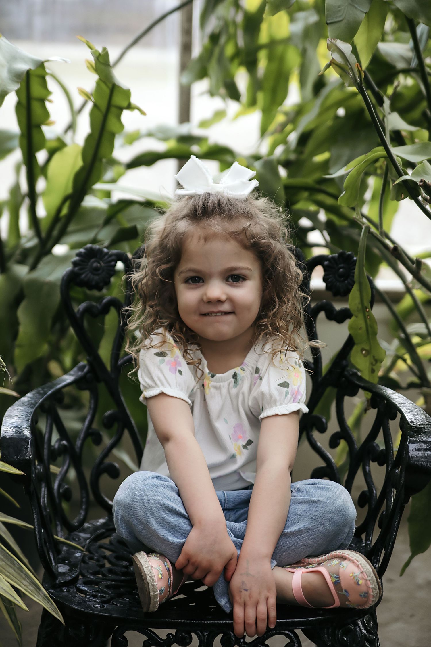 Curly-haired child sits cross-legged on a black wrought iron chair surrounded by lush green foliage.