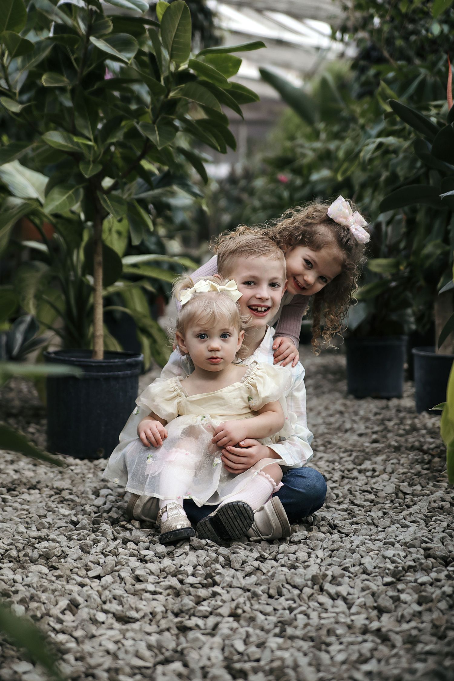 Two young children laugh and play together while sitting on gravel ground surrounded by potted plants in a greenhouse.