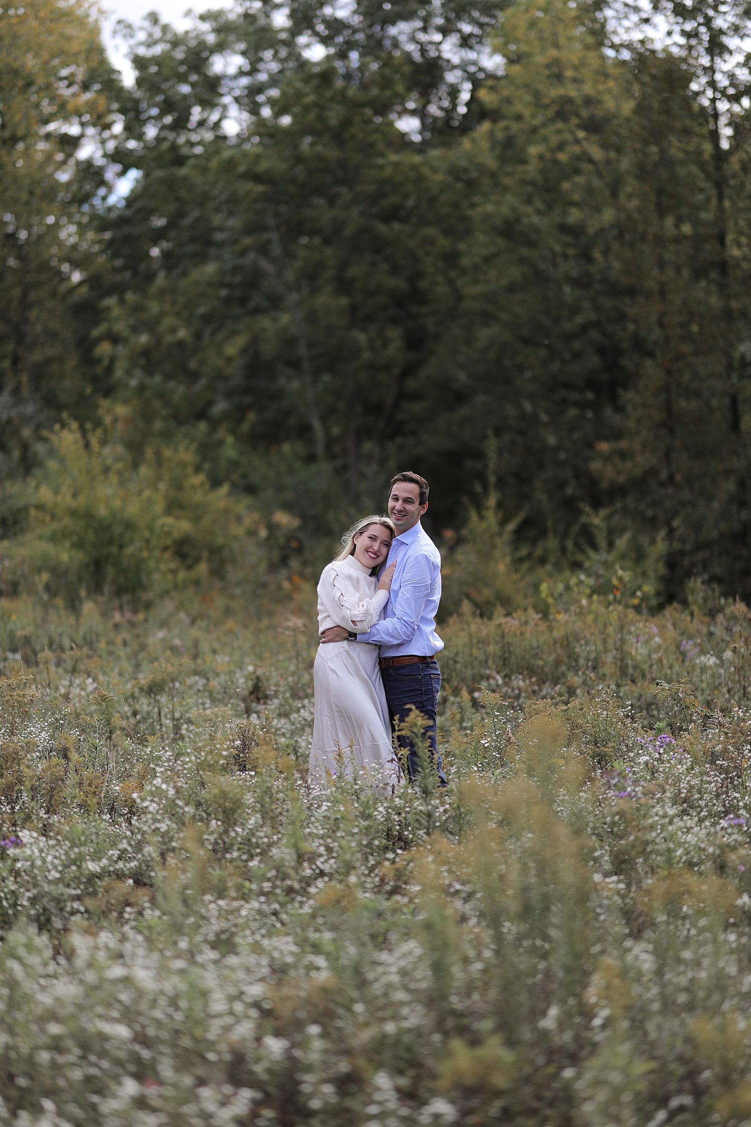 A couple embraces while standing in a field of wildflowers with trees in background.