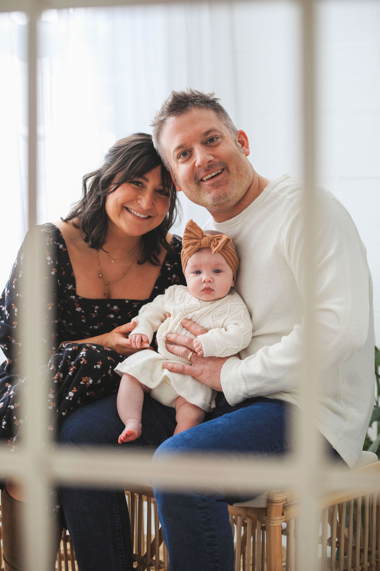 A family portrait shows a couple with their baby wearing a white dress and bow headband sitting together in a bright room.