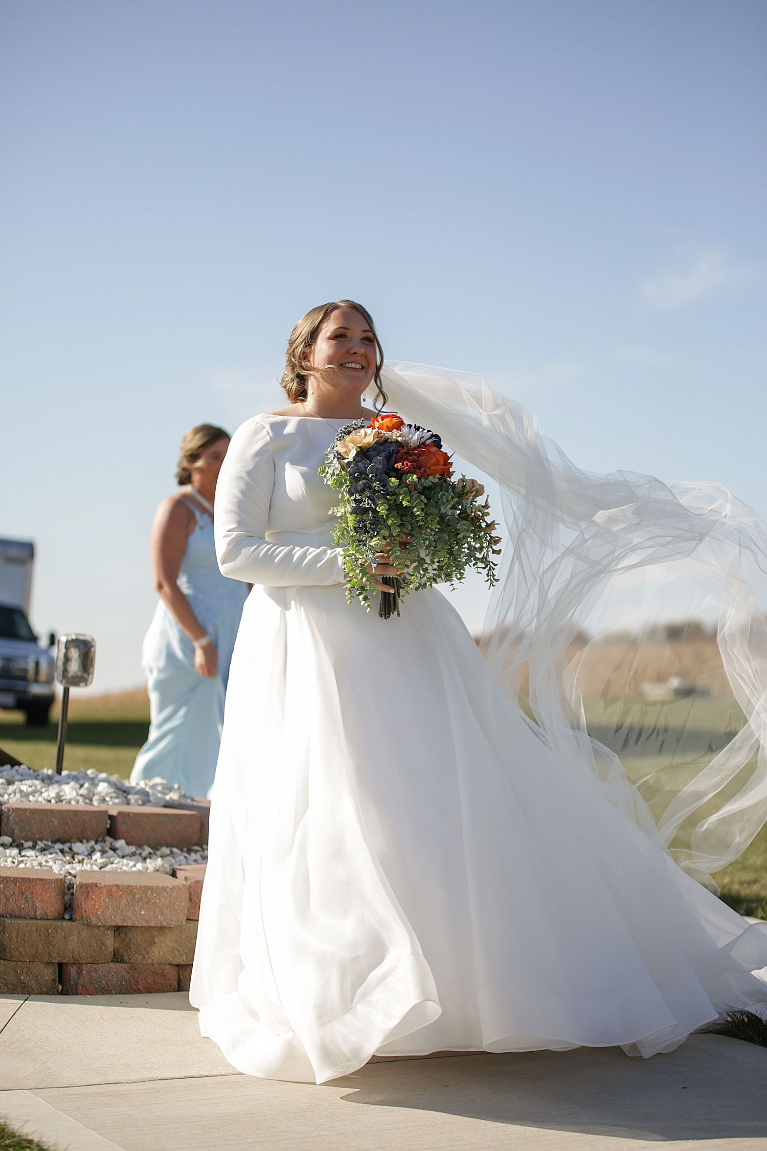 A bride in a long-sleeved white gown holds a colorful bouquet as her veil flows in the wind.
