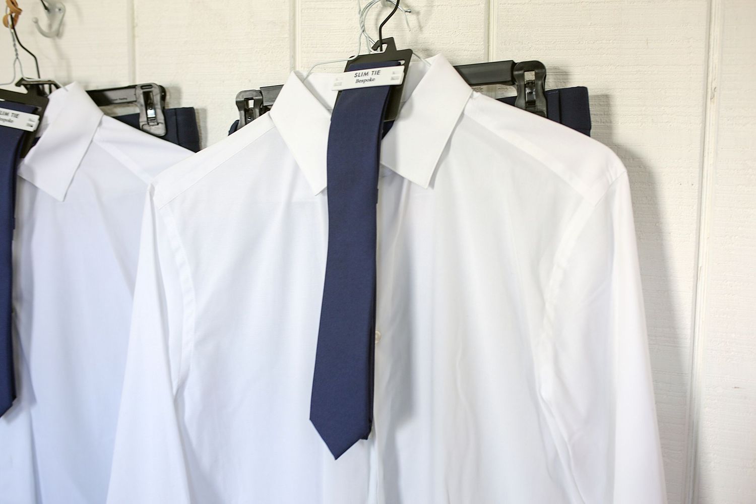 White dress shirts and a dark blue necktie hanging on hangers against a light colored wall.