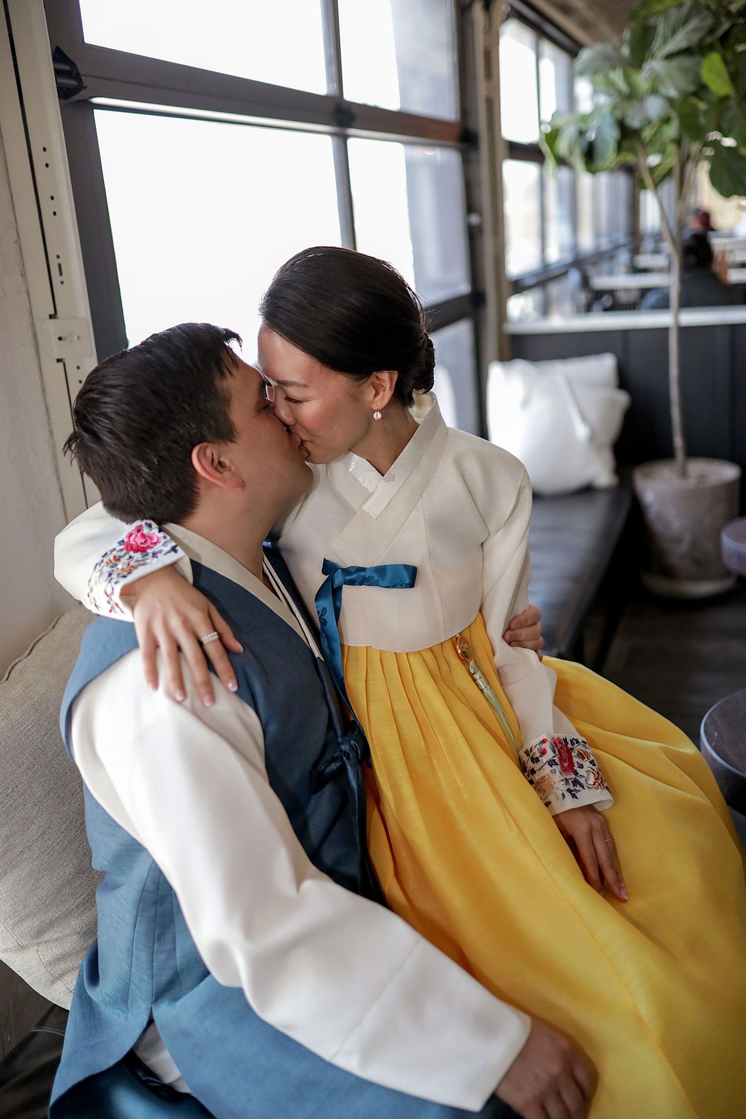 A couple in traditional Korean hanbok embrace and share tender moments in a bright sunlit corridor setting.