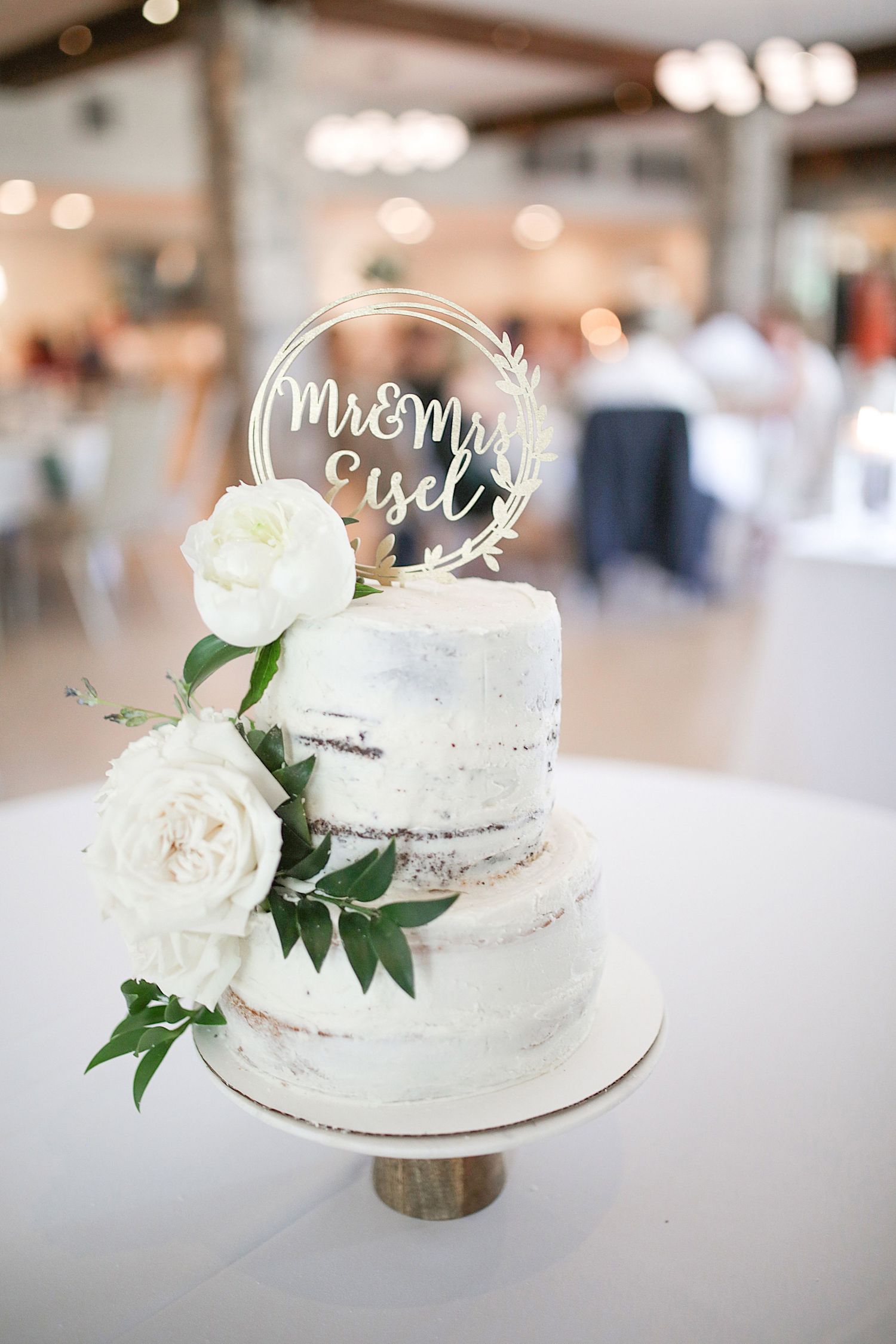 Elegant two-tier white wedding cake decorated with fresh roses and a circular 'Mr & Mrs' topper.