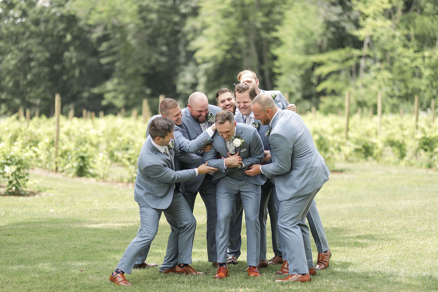 Groomsmen in matching grey suits huddle together playfully on a sunny lawn.