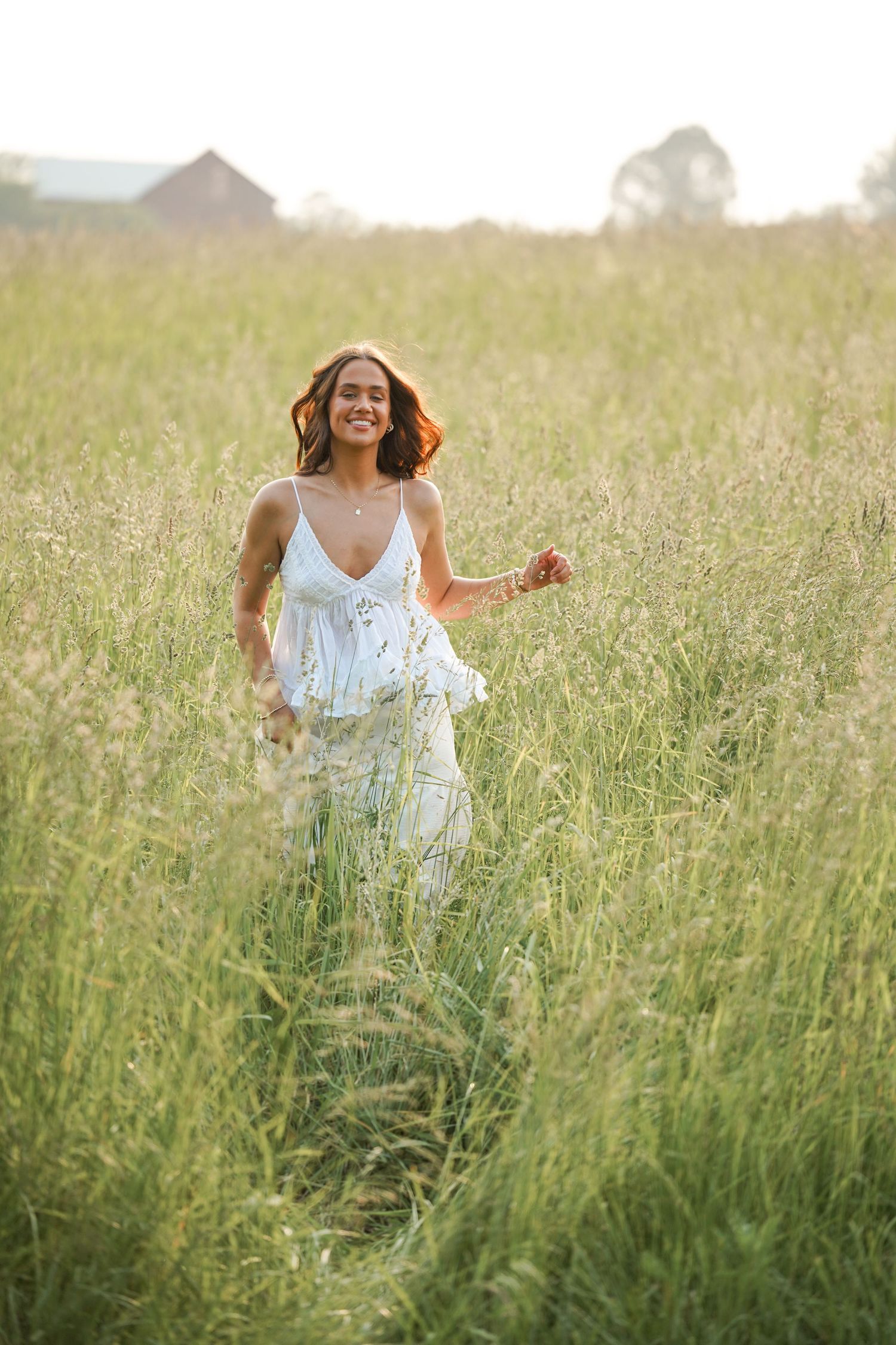 Someone in a white dress joyfully moves through a sunlit meadow of tall grasses at sunset.